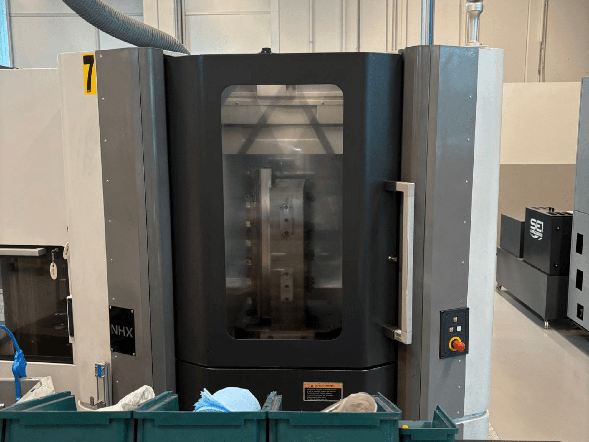 NHX CNC staklės iš priekio, matomas valdymo skydelis ir aiškus ekranas; staklės stovi dirbtuvėse, netoliese yra žaliosios dėžės.