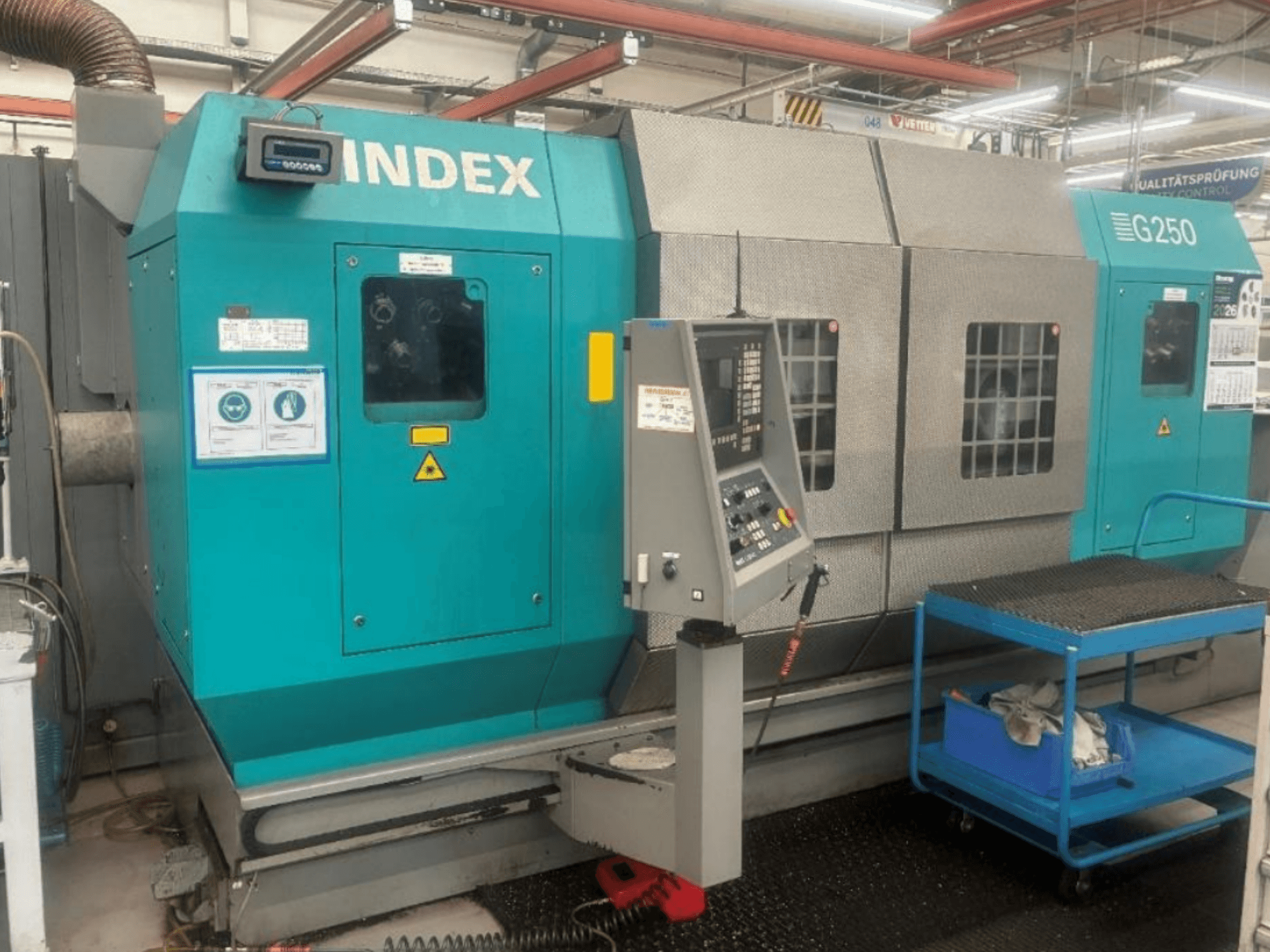Horizontali tekinimo staklė „INDEX G250“, vaizdas iš priekio, kuriame matomas valdymo skydas ir metalinis korpusas, su saugos etiketėmis ir mėlynu vežimėliu.