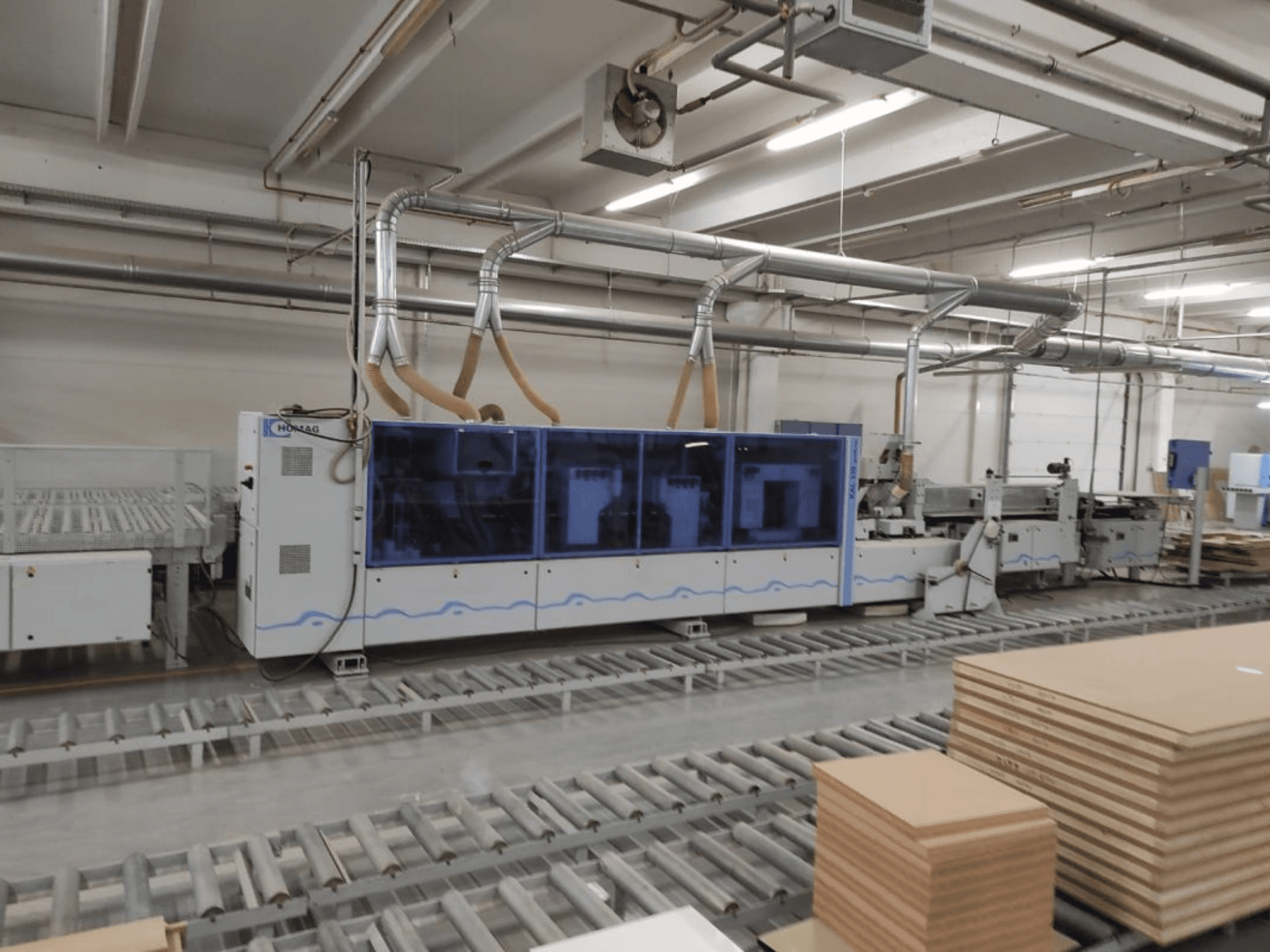 HOMAG gamybos mašina gamykloje, vaizdas iš priekio, mėlynas korpusas ir konvejerių sistema su sukrautais faneros lakštais.