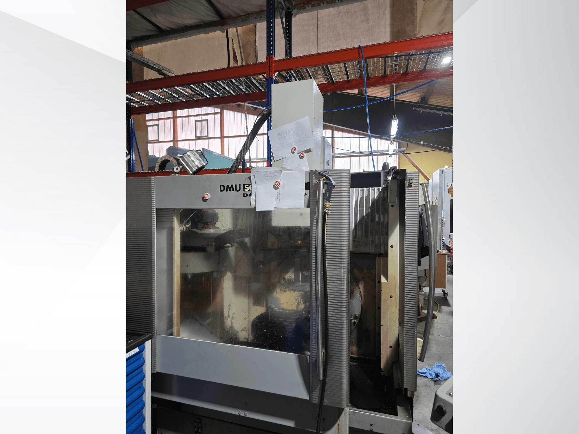 DMU 50 3 ašių CNC apdirbimo centras dirbtuvėse, parodytas iš priekio, su darbiniais komponentais ir pridėtais dokumentais.