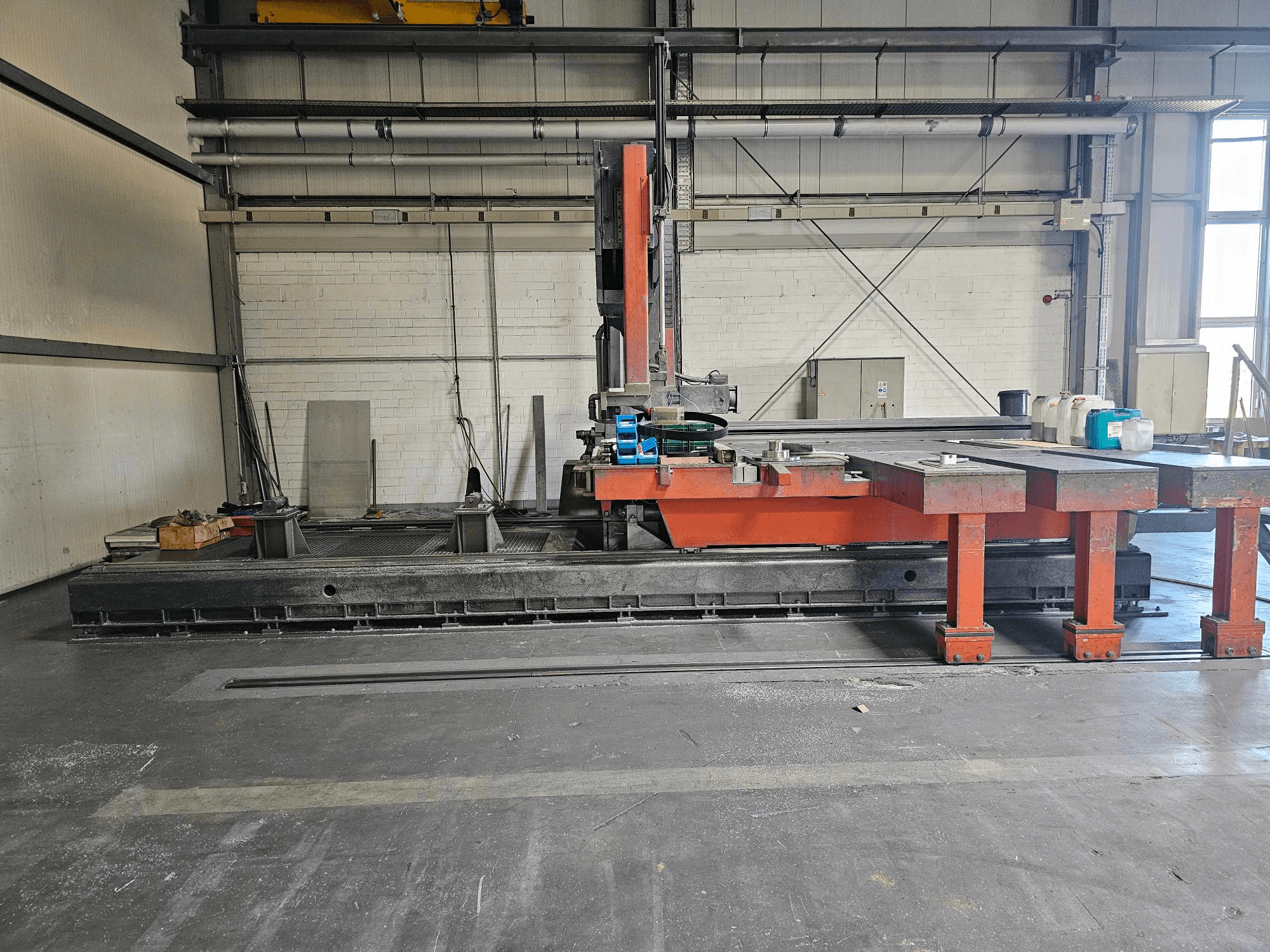 Pramoninės pjaustymo staklės atvirose dirbtuvėse, oranžinės ir juodos spalvos, su dideliu stalu; šoninis vaizdas, kuriame matyti priedai ir darbo vieta.