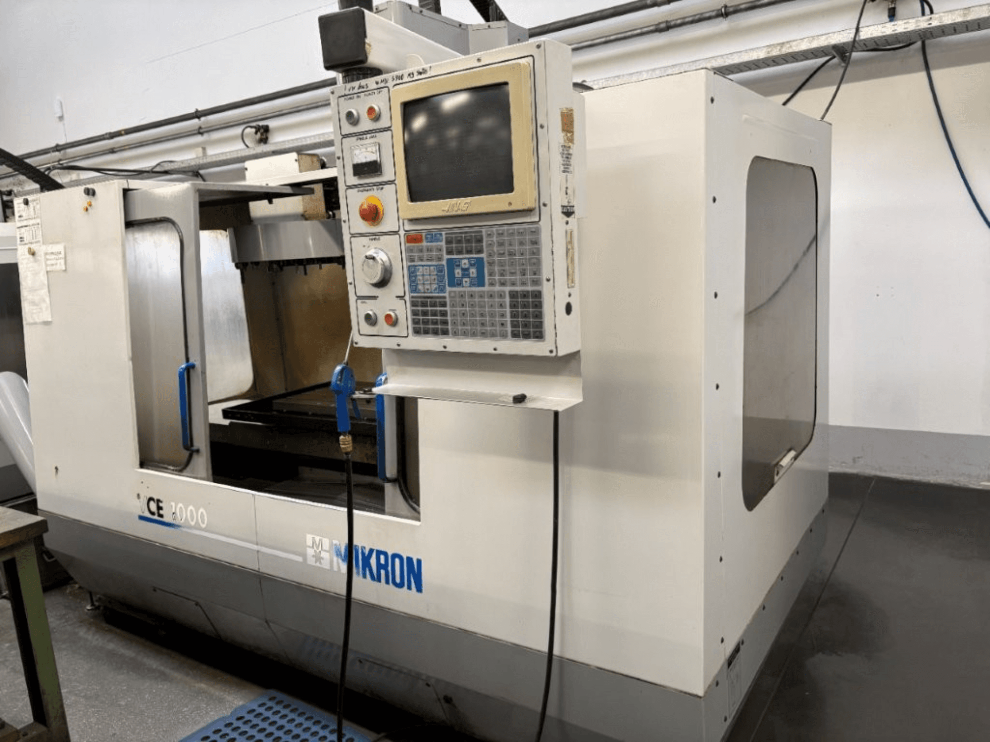 Mikron CE 2000 CNC staklės, vaizdas iš priekio; valdymo skydelis su mygtukais ir ekranu, sidabrinė išorė ir darbo zona.