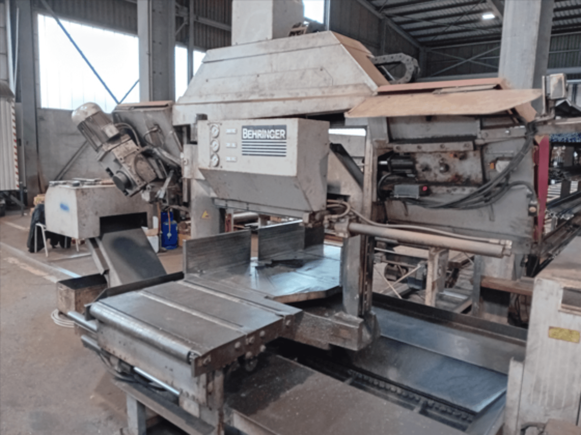 "Behringer" juostinis pjūklas, vaizdas iš priekio, turi didelį diską, variklį ir reguliuojamą stalą, idealiai tinka metalo pjovimui dirbtuvėse.