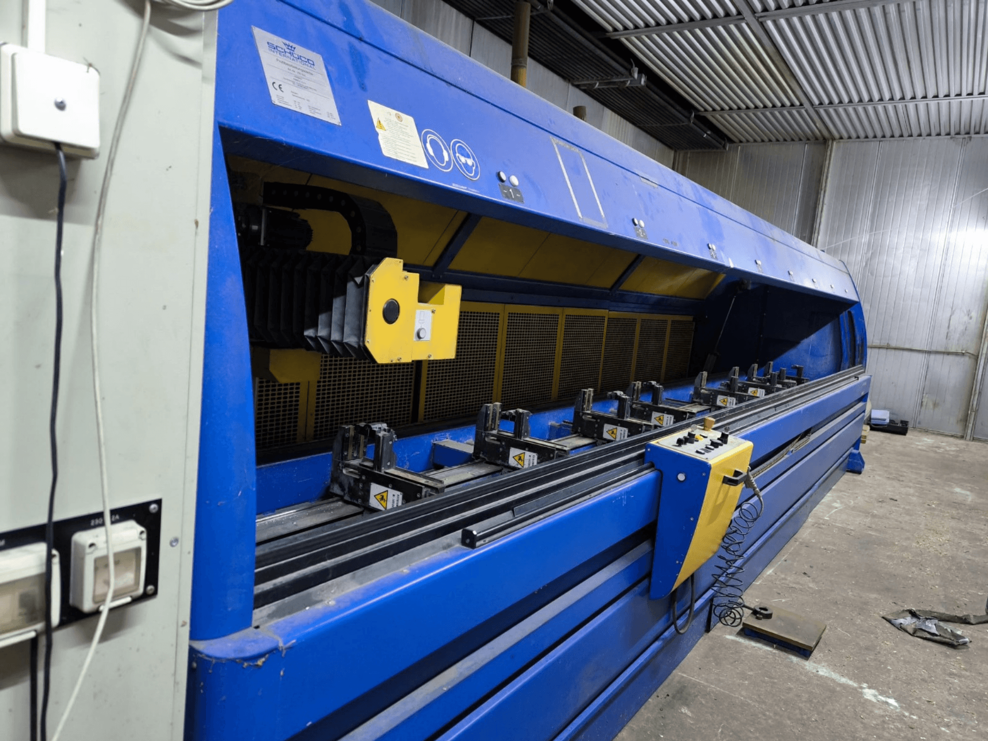 Mėlynos spalvos "Schneider" pramoninės pjaustymo staklės, vaizdas iš šono, matomas valdymo skydelis, diržai ir kelios prispaudimo stotys.