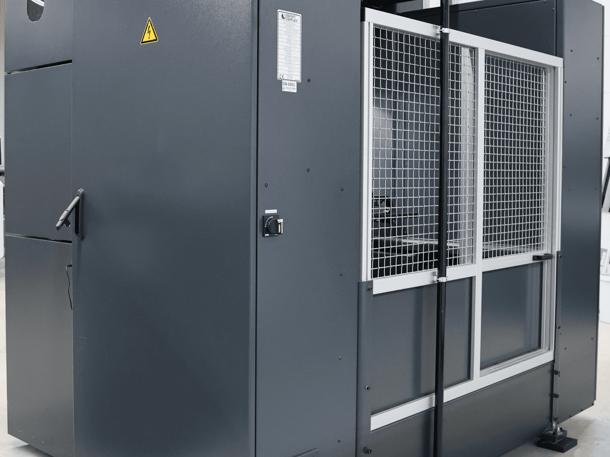 Pilka pramoninė mašina su saugos įspėjimu, priekinis vaizdas, kuriame matomos tinklinės durelės ir valdymo skydelis, modelis paženklintas etikete.