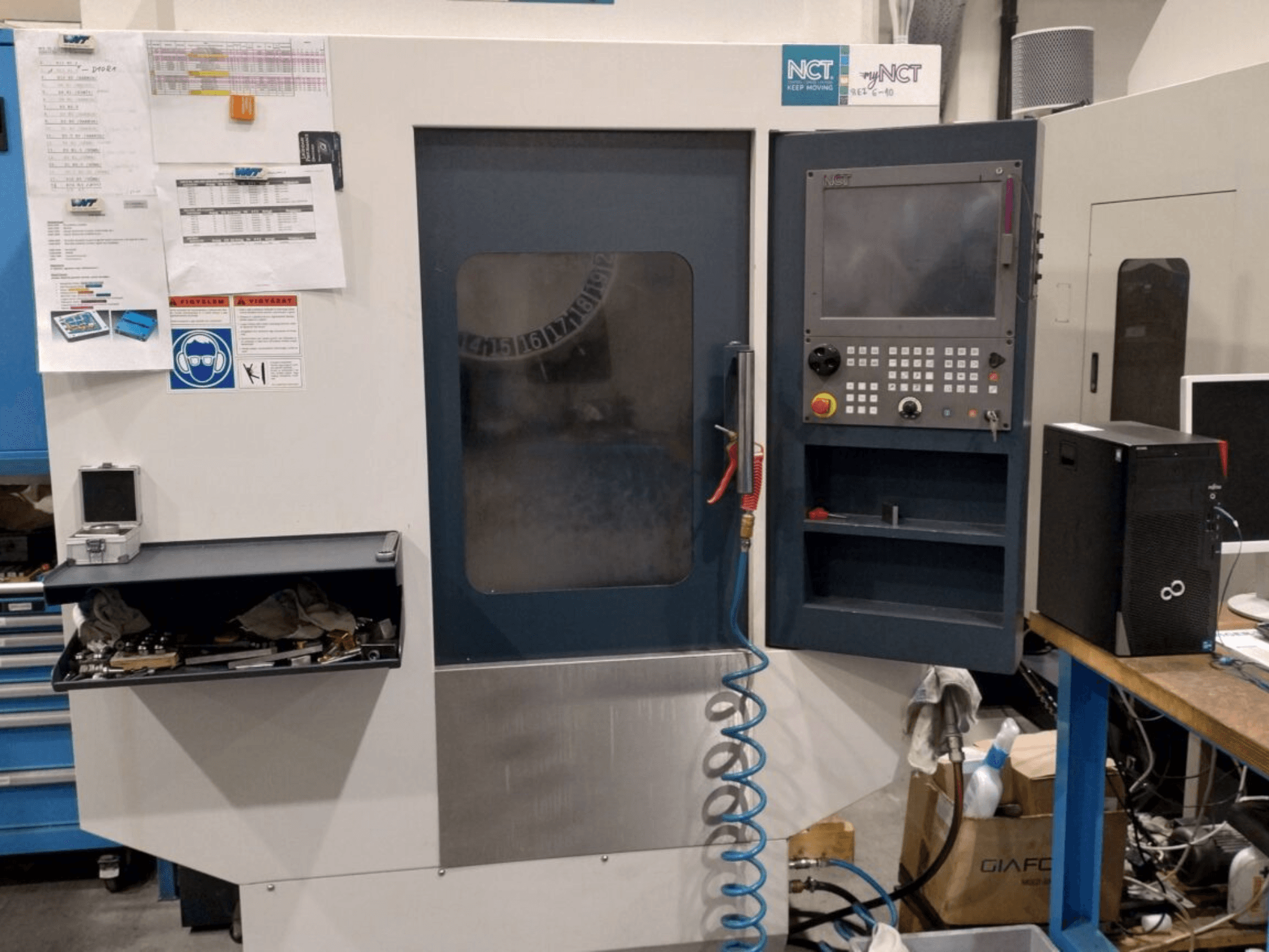 NCT CNC staklės, vaizdas iš priekio, valdymo skydelis su skaitmenine klaviatūra, įrankiai laikymo stalčiuje ir instrukcija ant sienos.