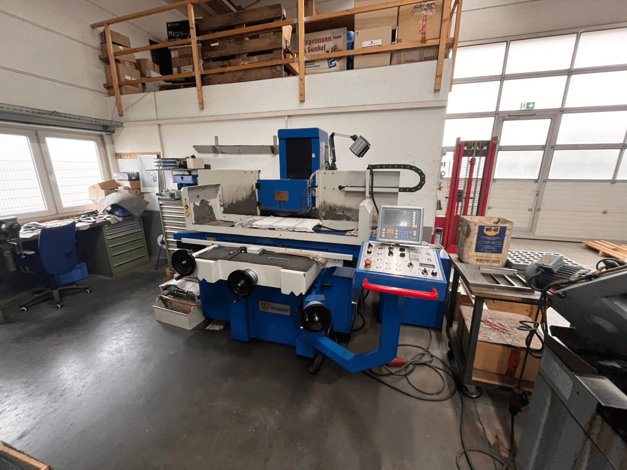 Mėlyna paviršiaus šlifavimo mašina su skaitmeniniu valdymo skydeliu, vaizdas iš priekio, matoma darbo vietos netvarka ir žalia įrankių spintelė.