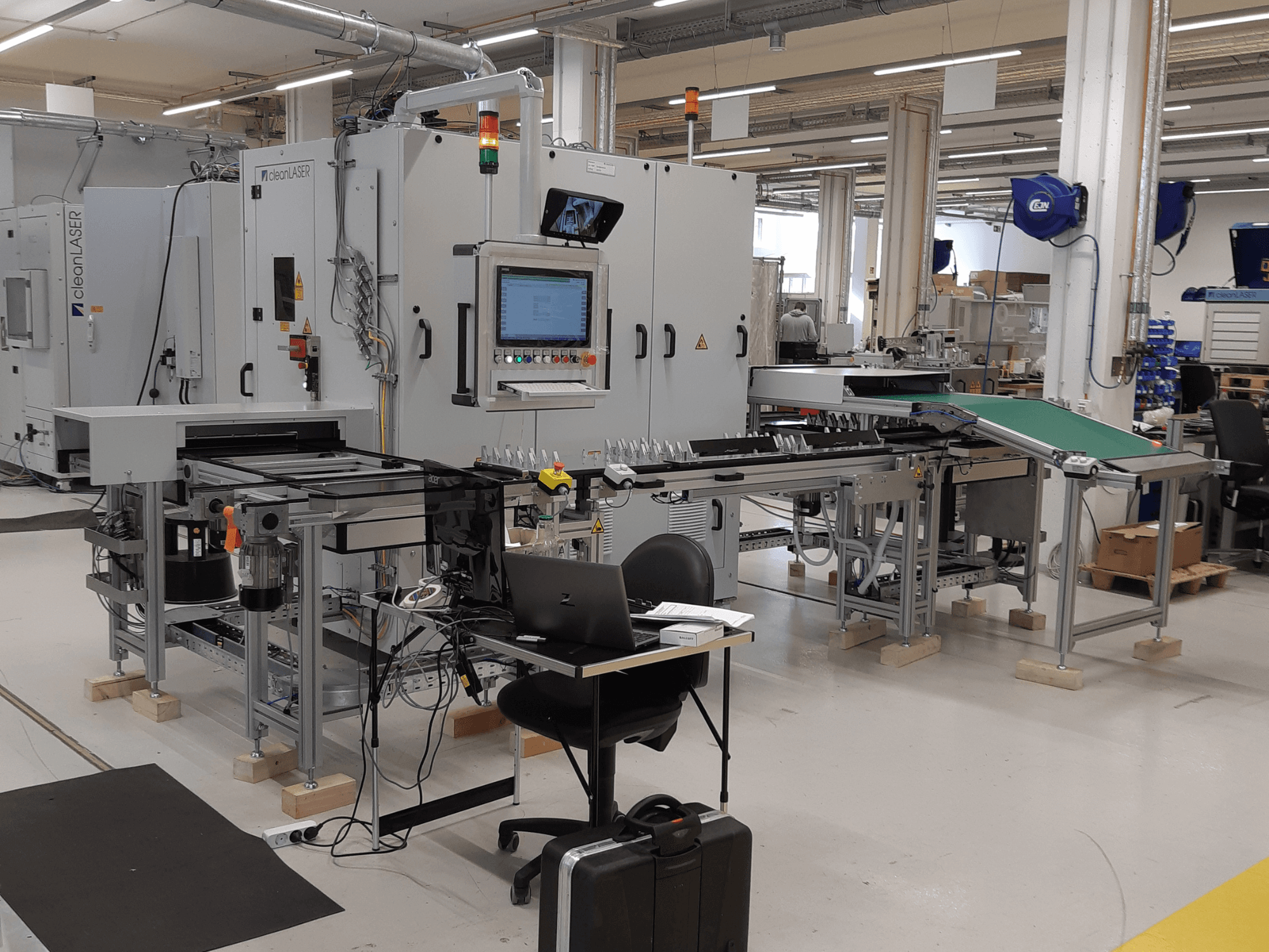 „CLEAN LASER“ staklės „clean CELL 3220“, vaizdas iš priekio, kuriame matomas valdymo pultas, konvejeris ir darbo vietos išdėstymas gamybinėje aplinkoje.