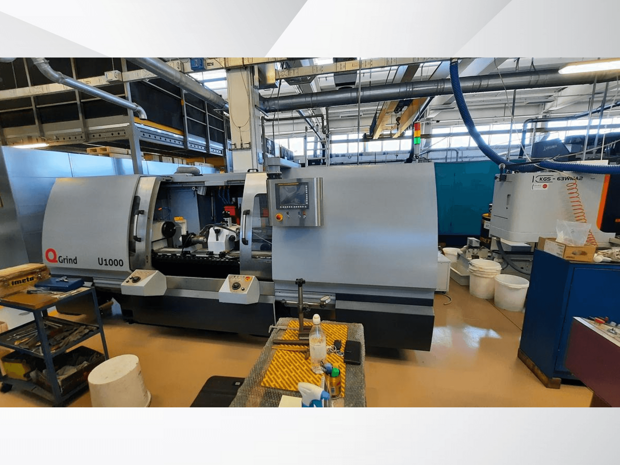 "Q-Grind U1000" staklės dirbtuvėse, kuriose matomas priekinis vaizdas su valdymo skydeliu ir atvira pjovimo zona, apsupta įrangos.