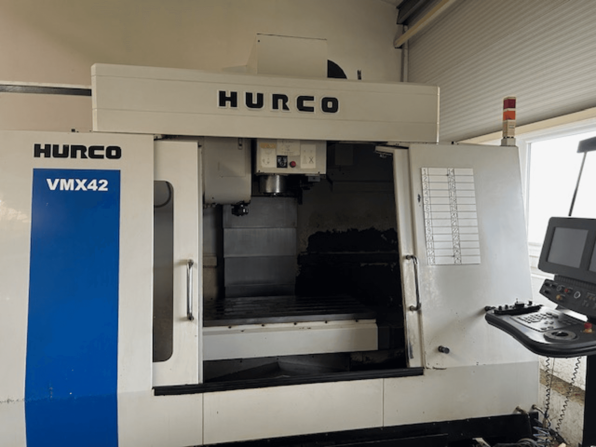 Hurco VMX42 CNC staklių vaizdas iš priekio, kuriame matyti valdymo skydelis, suklys ir darbo zona; atskleidžiamas darbinis išdėstymas.