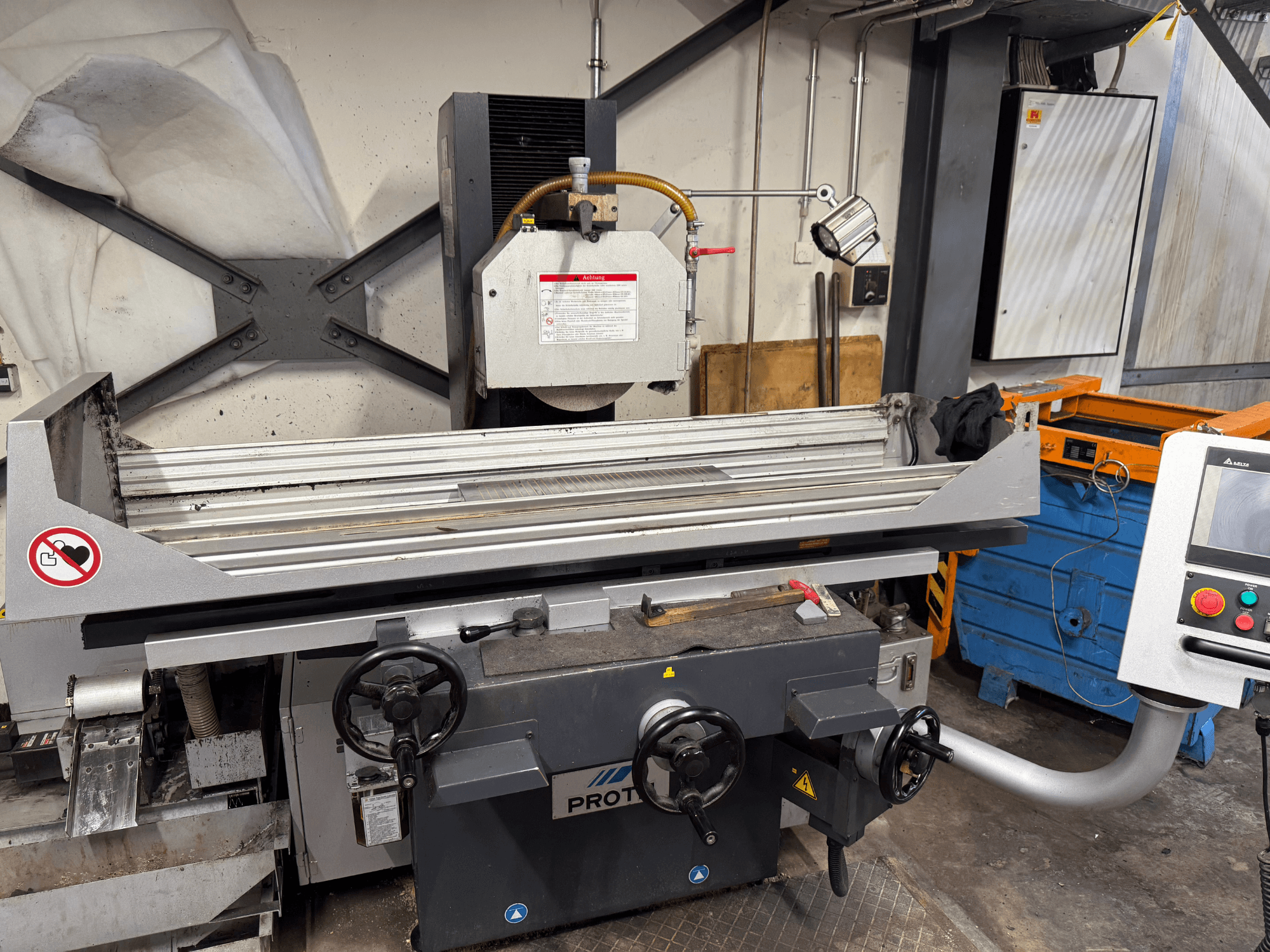 PROTEC mašinos priekinis vaizdas, kuriame matyti paviršiaus šlifuoklis su rankiniu valdymu, saugos įspėjimais ir aušinimo sistema.