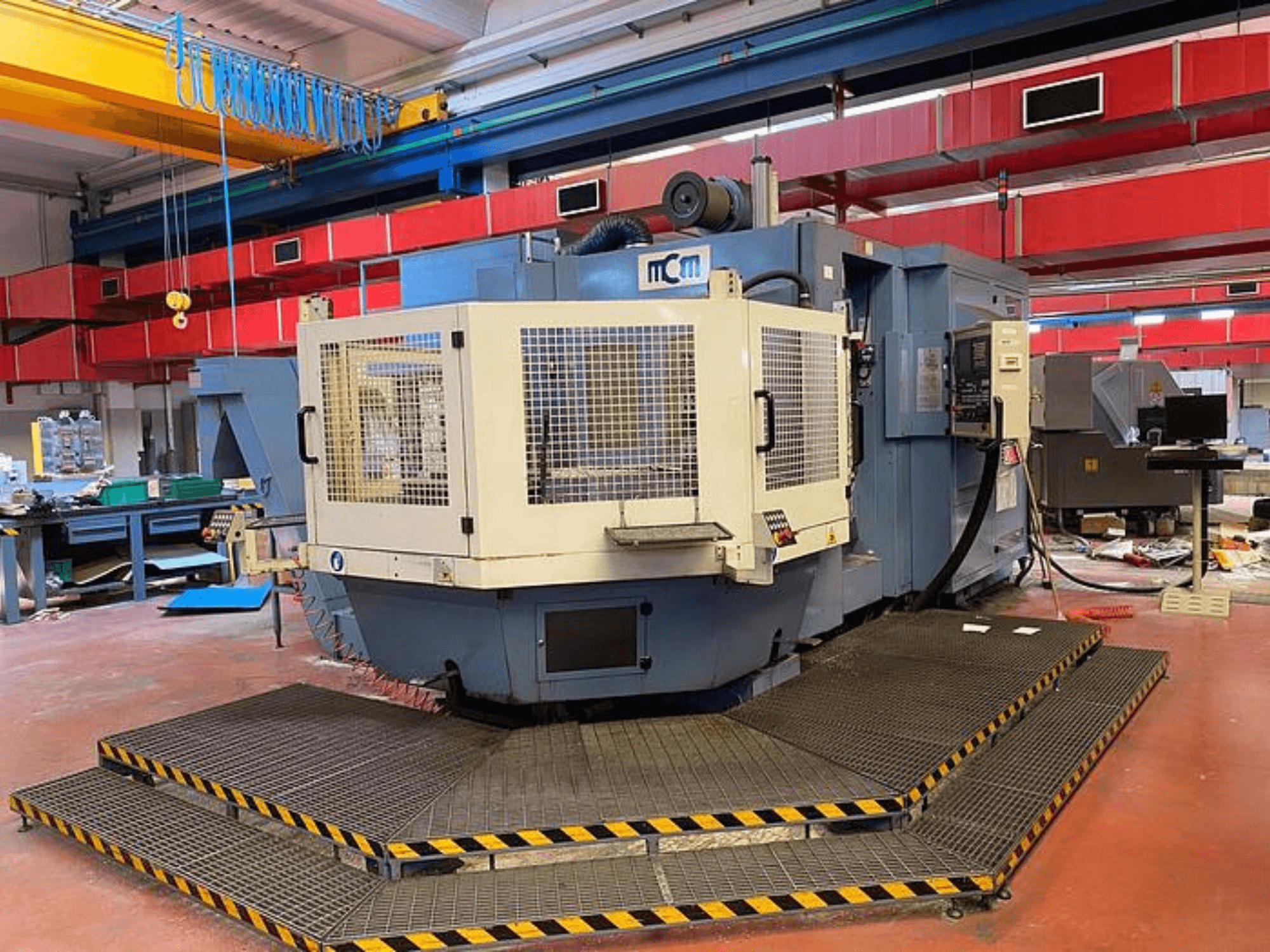 MCM CNC staklės gamykloje, vaizdas iš priekio su apsauginiu narveliu, juodais ir geltonais apsauginiais kraštais, apsuptas pramoninės įrangos.