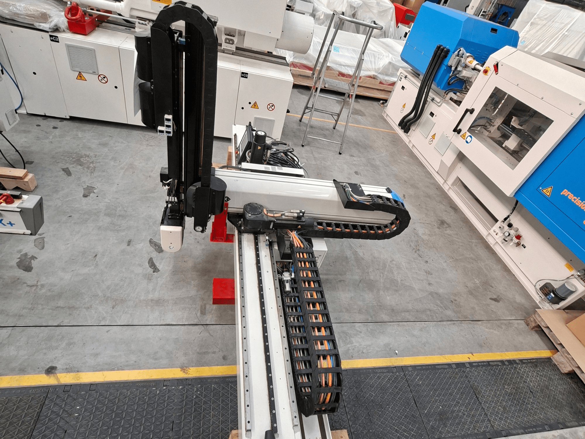Plastiko perdirbimo staklės „Krauss Maffei LRX150-MC5“, vaizduojamos iš viršaus, kuriose matoma robotinė ranka ir kabelių trasos gamybos salėje.