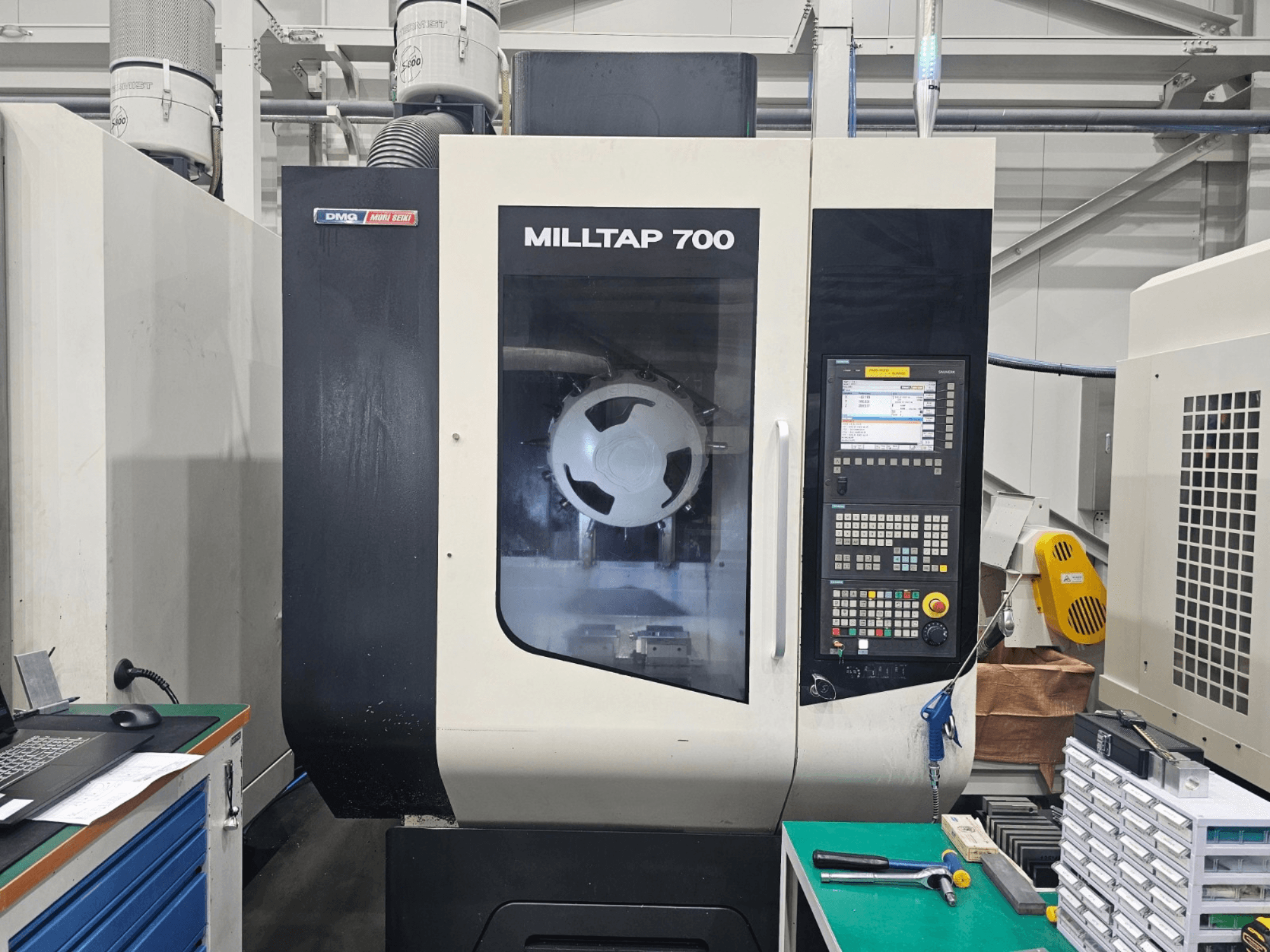 Vertikalusis apdirbimo centras „DMG MORI MILLTAP 700“, vaizdas iš priekio, kuriame matomi valdymo skydas, suklis ir darbo erdvės detalės.
