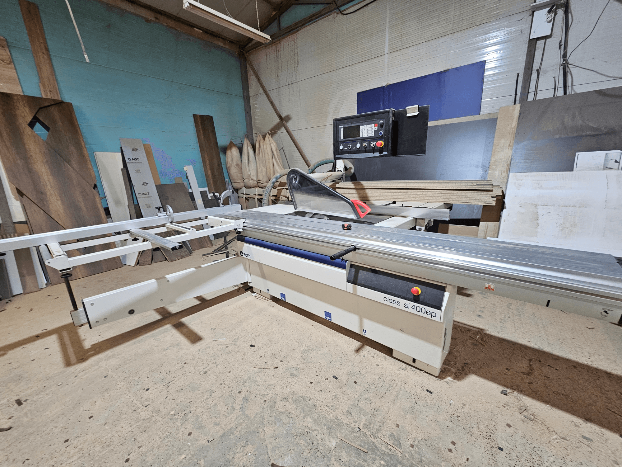 SI400EP klasės plokščių pjūklas, vaizdas iš priekio, su reguliuojama tvorele, pastatytas ant smėlio dirbtuvių grindų, apsuptas medienos plokščių.