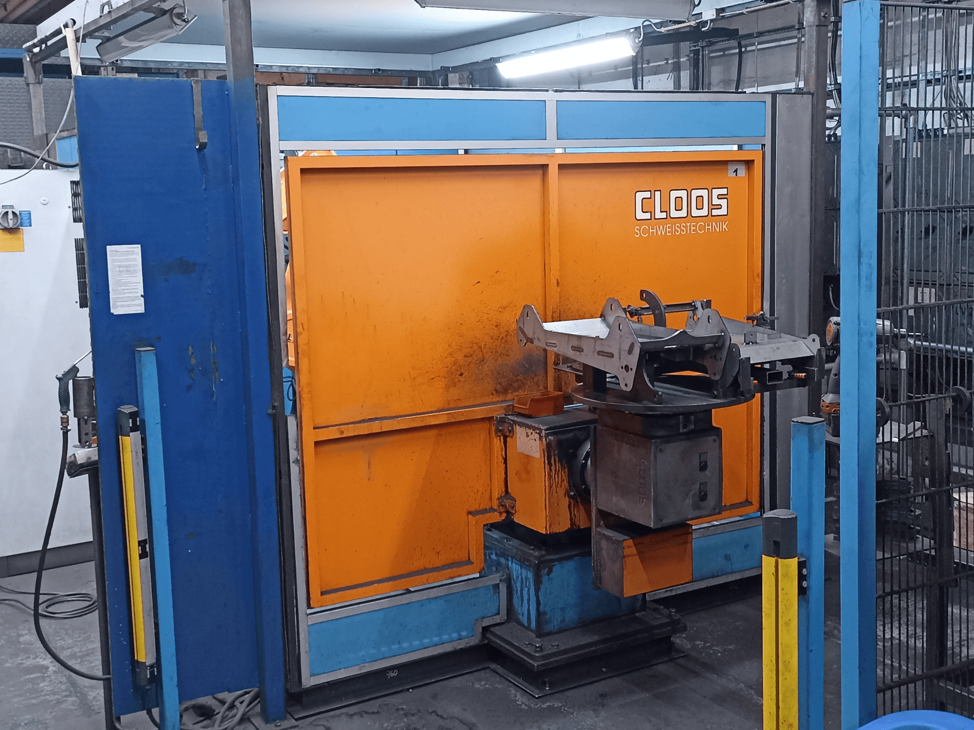 CLOOS „Romat 320“ suvirinimo robotas dirbtuvėse, vaizdas iš priekio, su oranžiniu valdymo pultu ir metaliniu ruošiniu ant besisukančio pagrindo.
