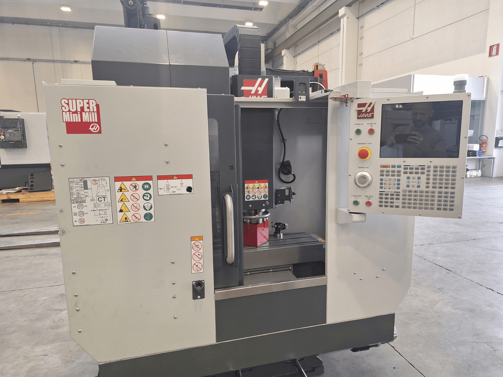 „HAAS Super Mini Mill“ vaizdas iš priekio, kuriame matomas valdymo skydas, saugos etiketės ir apdirbimo zona su įtvirtintu ruošiniu.