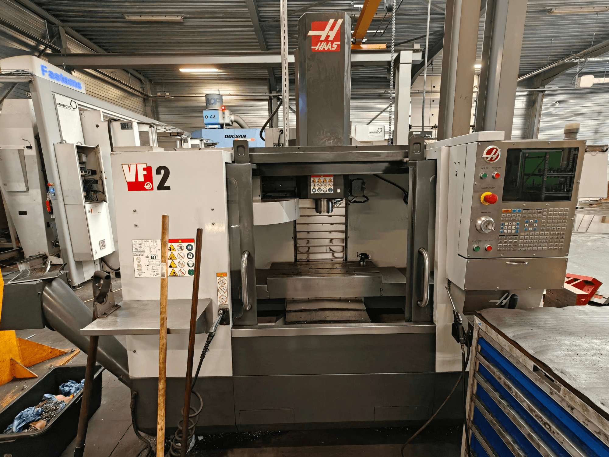 Vertikalusis apdirbimo centras „HAAS VF-2“, vaizdas iš priekio, kuriame matomi valdymo elementai, suklis ir darbo stalas dirbtuvės aplinkoje.