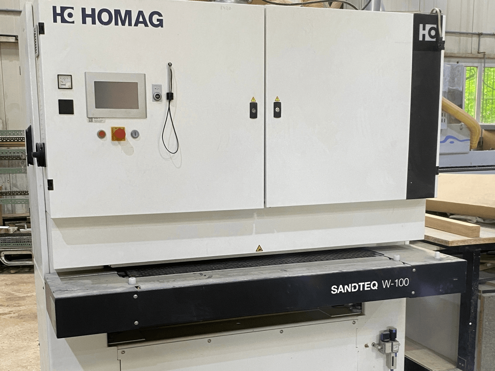 „HOMAG SANDTEQ W-100“ juostinis šlifuoklis dirbtuvėse: matomas valdymo skydelis, dulkių išsiurbimo anga ir šlifavimo juostos sritis. Vaizdas iš priekio.