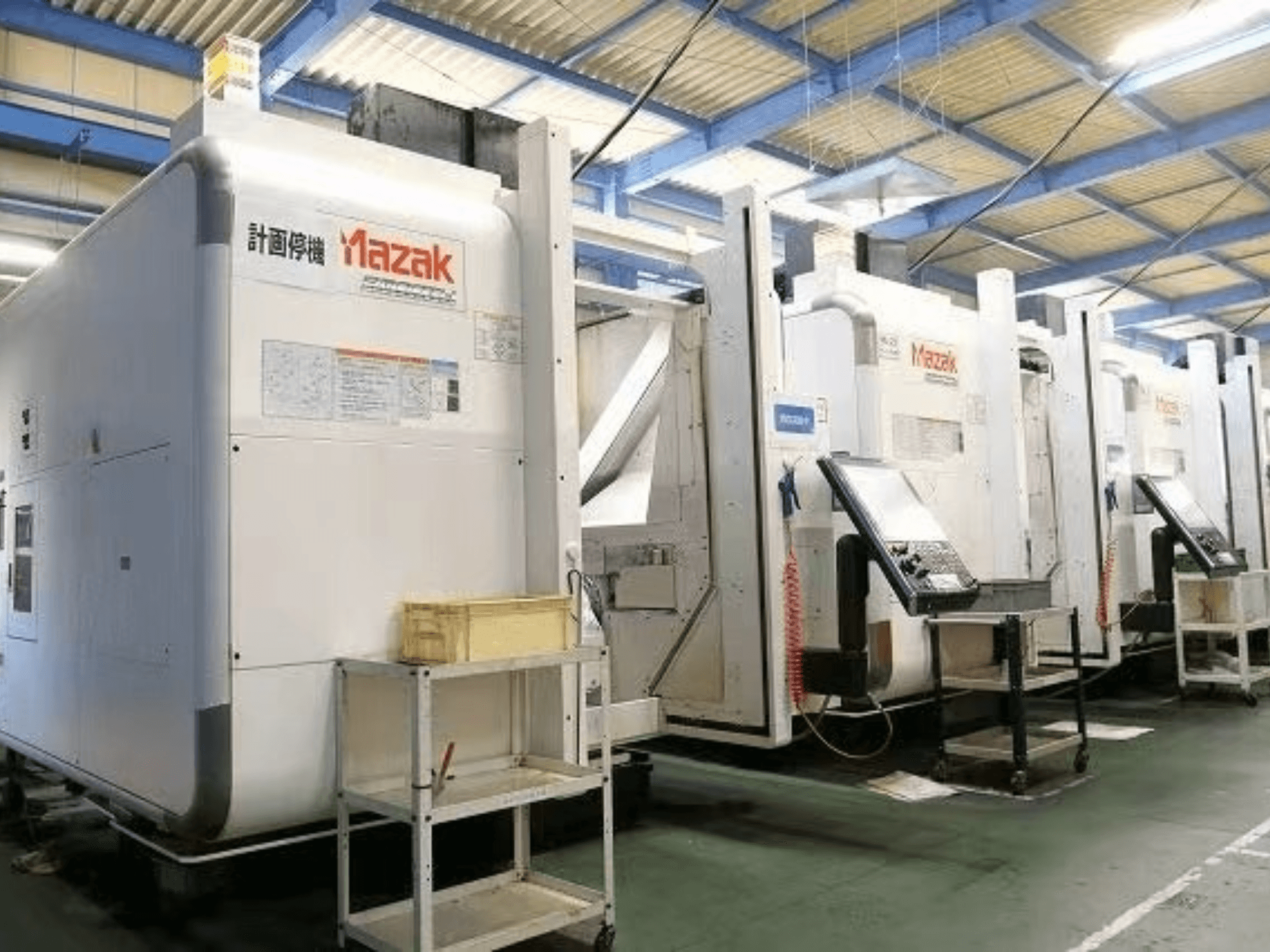 "Mazak" CNC staklės gamykloje, šoninis vaizdas, kuriame matomi valdymo skydai ir etiketės ant baltų korpusų.