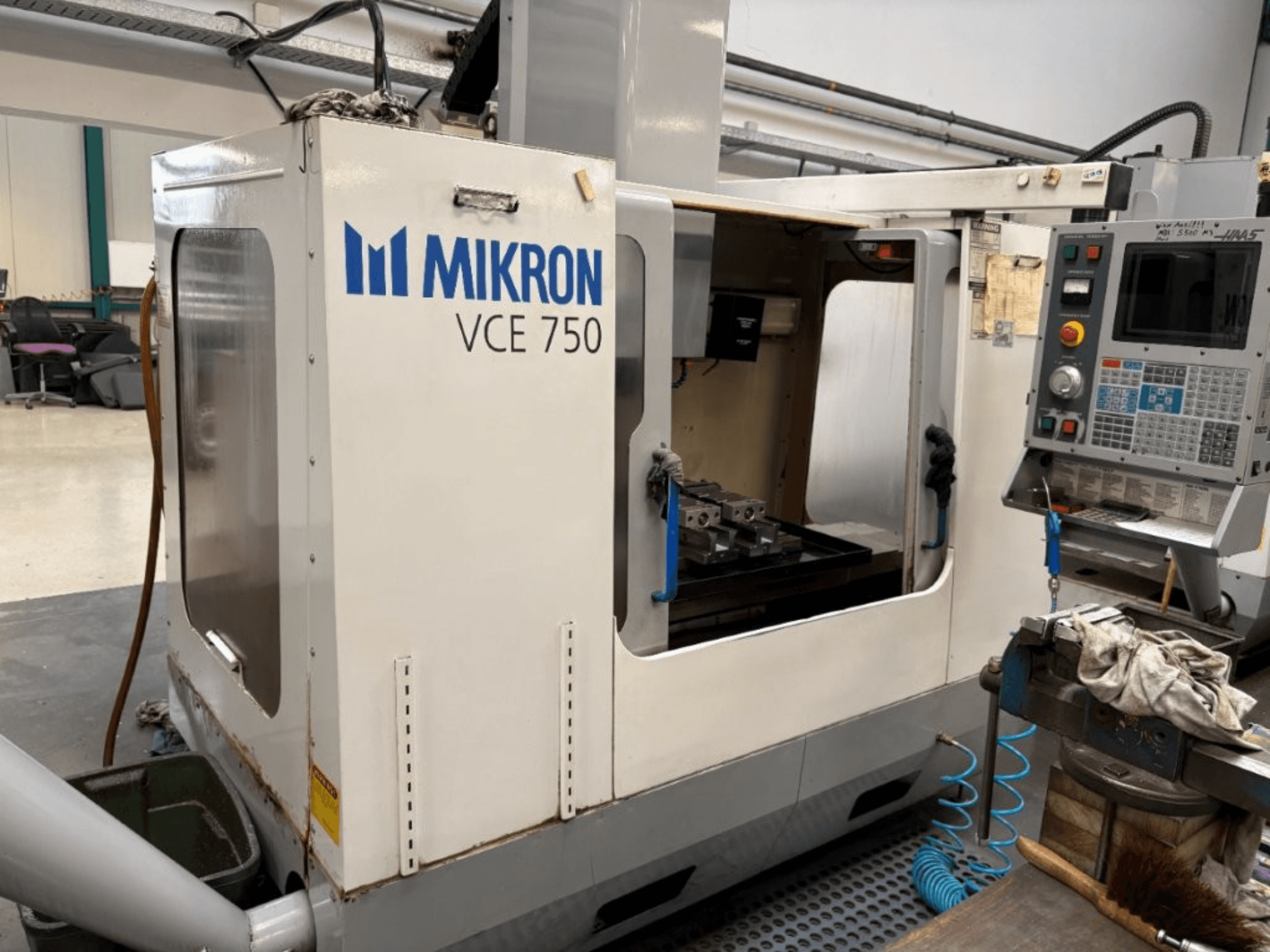 Mikron VCE 750 CNC frezavimo staklių priekinis vaizdas, kuriame matomas apdirbtas stalas ir valdymo skydelis su įvairiais mygtukais ir ekranais.
