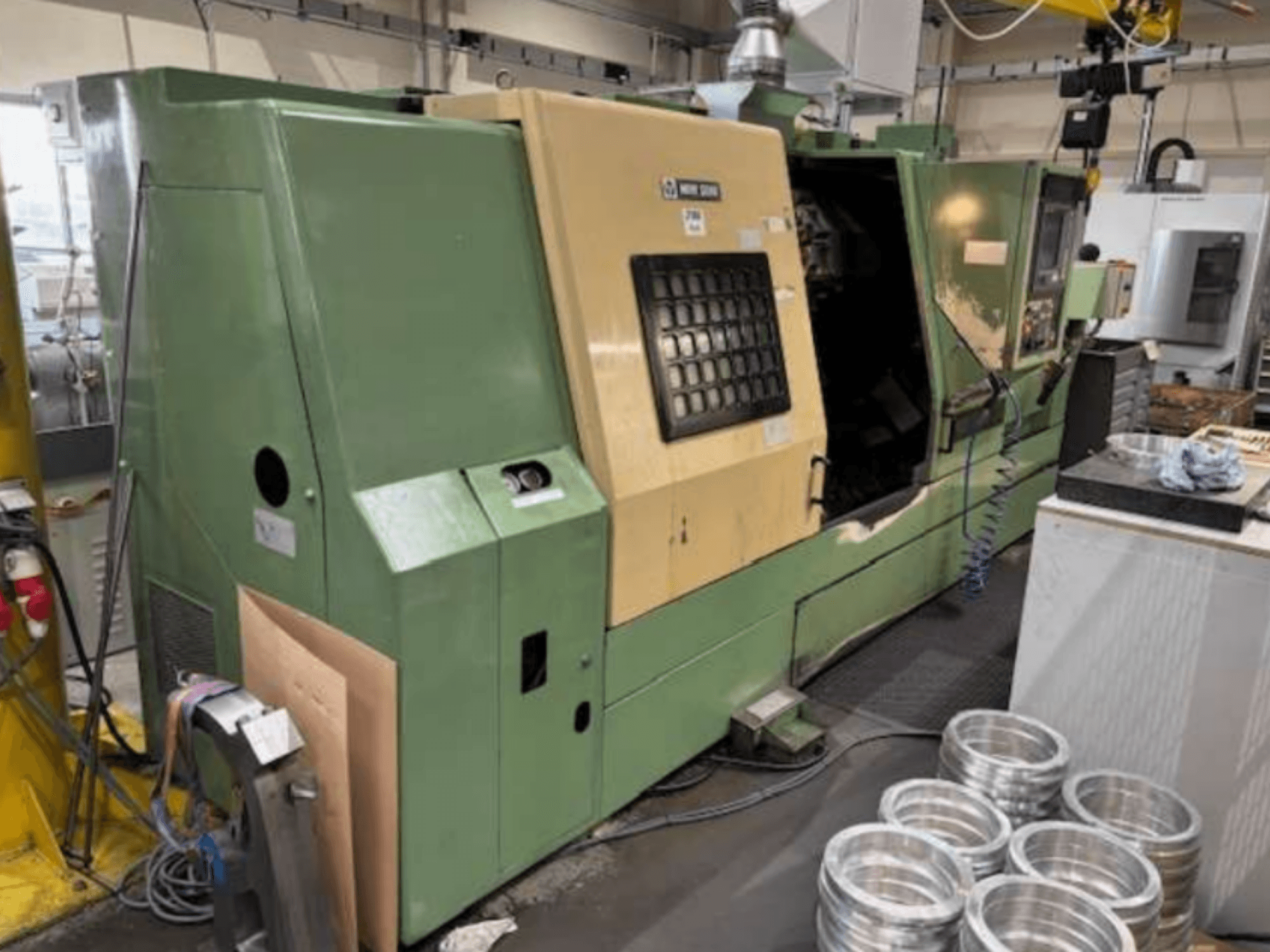 Žalios "Mazak" CNC tekinimo staklės, žiūrint iš priekio, su valdymo pultu ir apdirbamomis aliuminio detalėmis ant darbastalio.