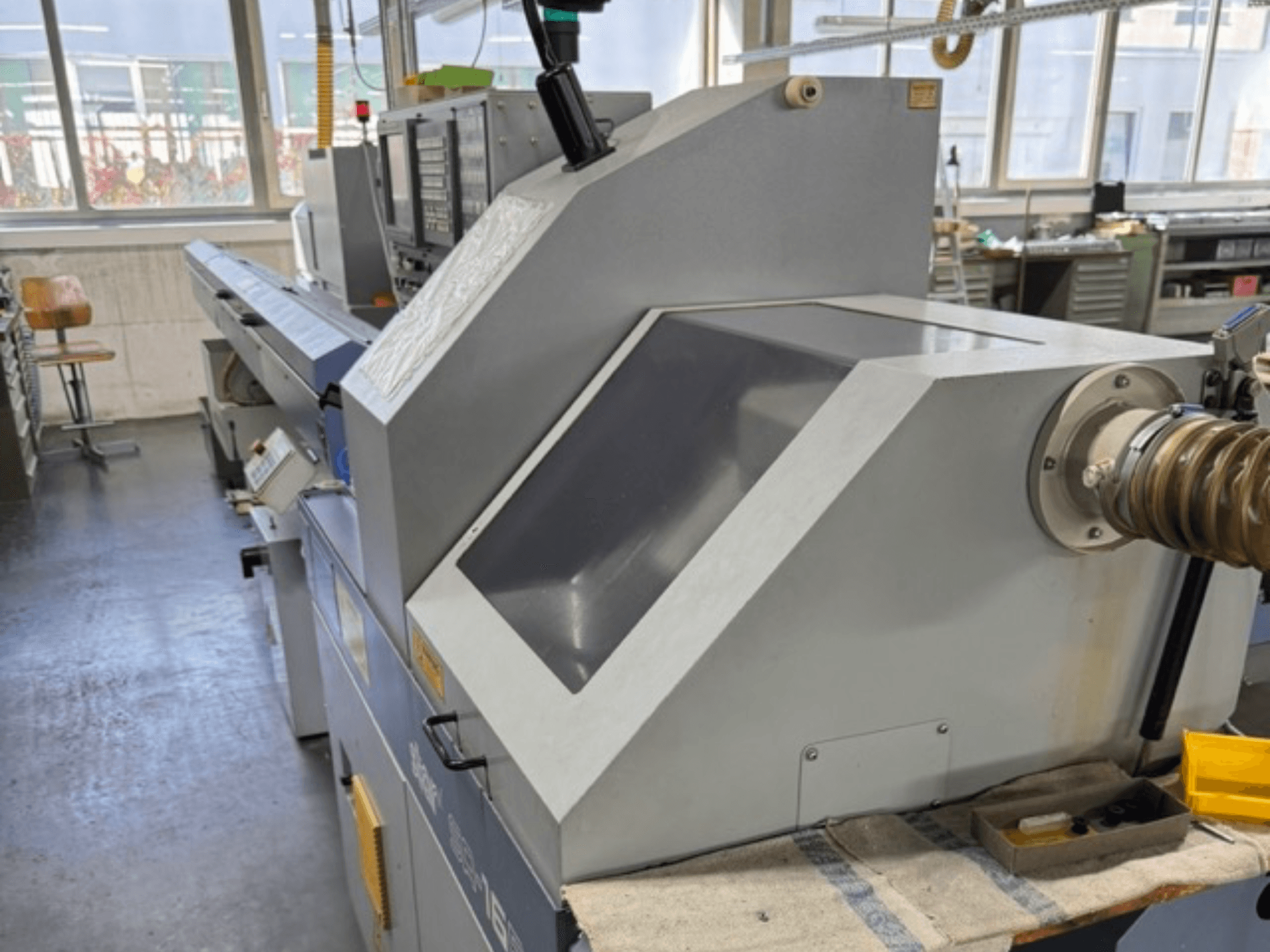 Pilka pramoninė mašina, žiūrint iš šono, turi didelį kampinį skydelį, valdymo mygtukus ir žarnos priedą.