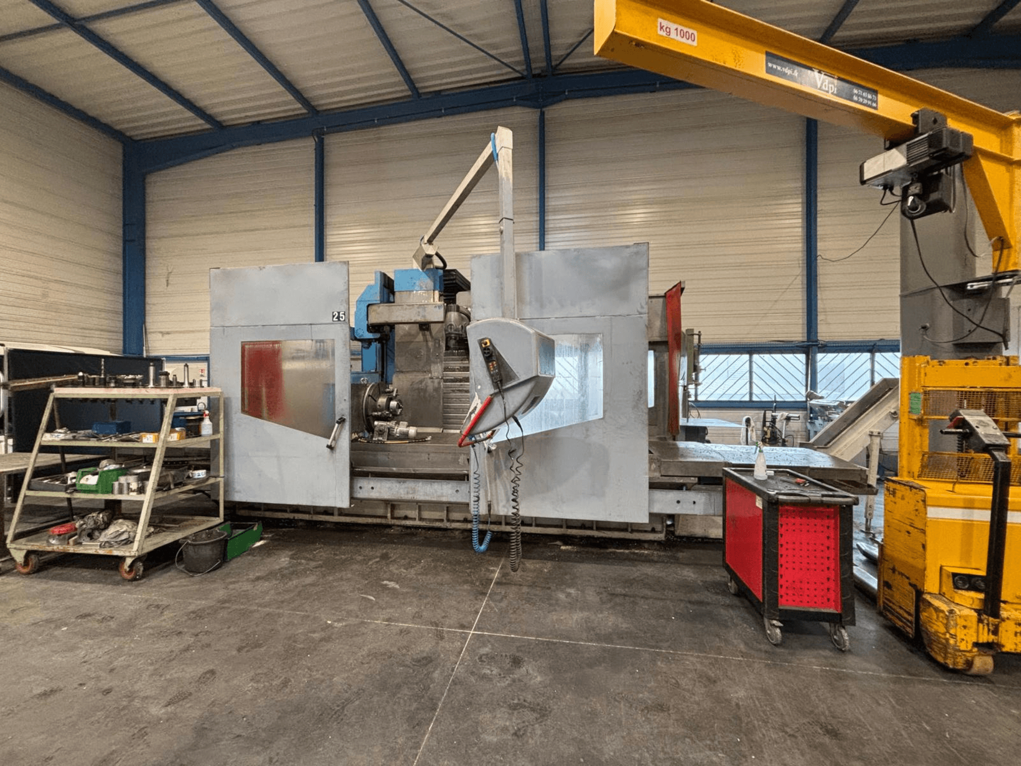 CNC frezavimo staklės dirbtuvėse iš šono; matomi įrankiai ir laikymo lentynos. Prekės ženklas ir modelis nenurodyti.