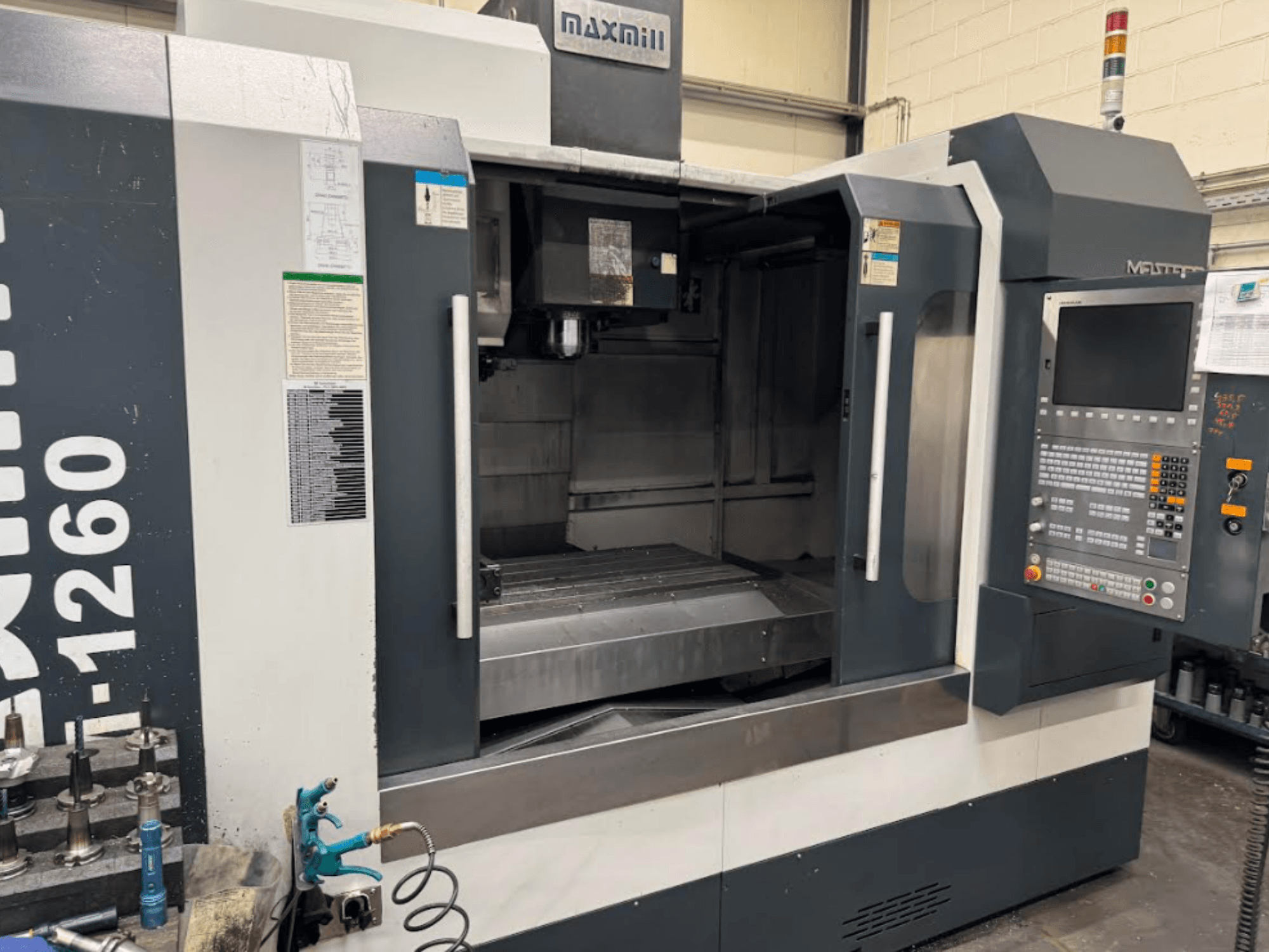 Maxmill CNC frezavimo staklės, modelis M-1260, priekinis vaizdas, kuriame matyti tuščia darbo vieta su valdymo skydeliu ir naudojimo instrukcijomis.