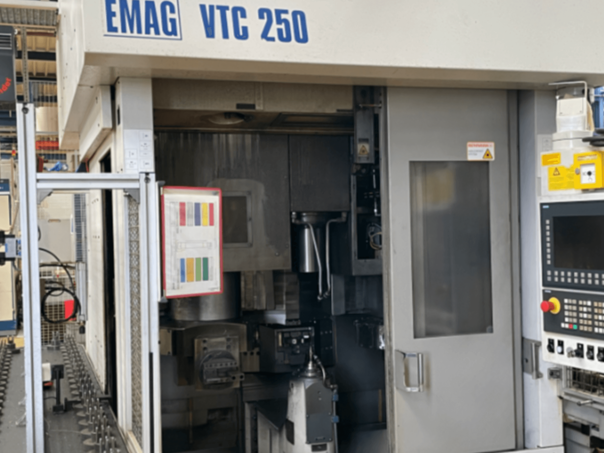 EMAG VTC 250 vertikalios tekinimo staklių vaizdas iš priekio, kuriame matomas valdymo skydas, darbo sąsaja ir atidarytos prieigos durys.
