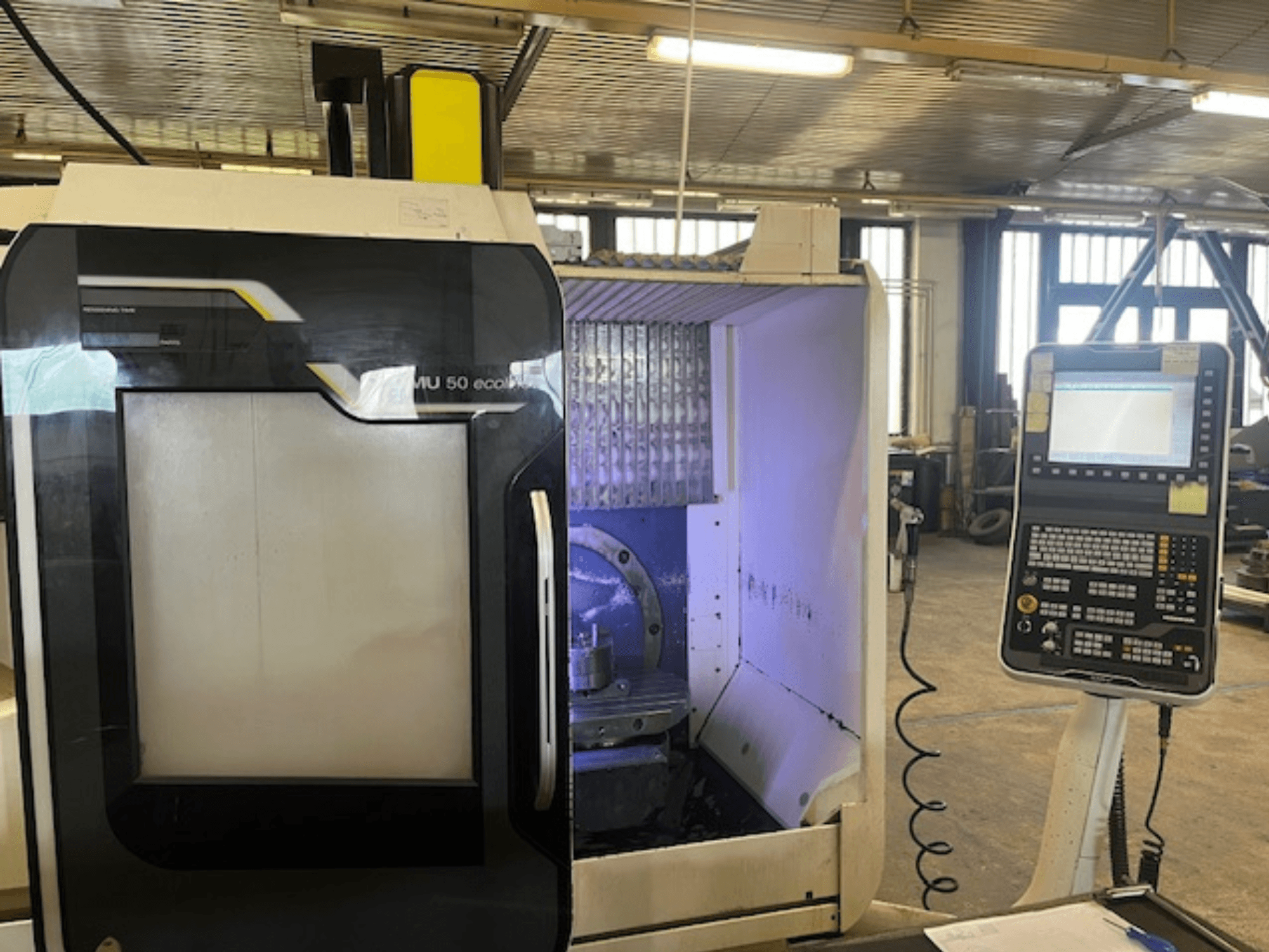 DMU 50 eco frezavimo staklių priekinis vaizdas su valdymo skydeliu, įrankių laikikliu ir darbo vieta.