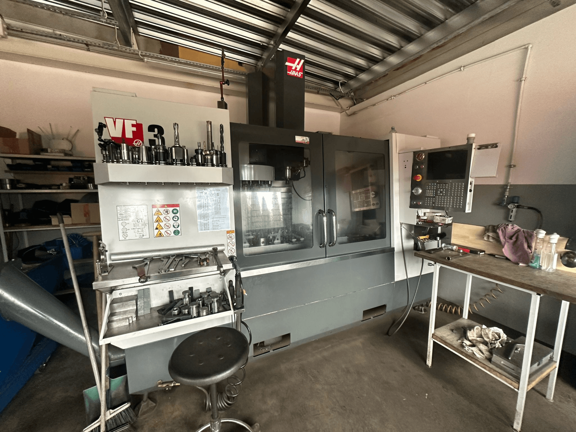 Haas VF-3 CNC staklės dirbtuvėse, vaizdas iš priekio su įrankiais ir valdikliais ant gretimo stalo, rodoma techninė sąsaja ir saugos informacija.