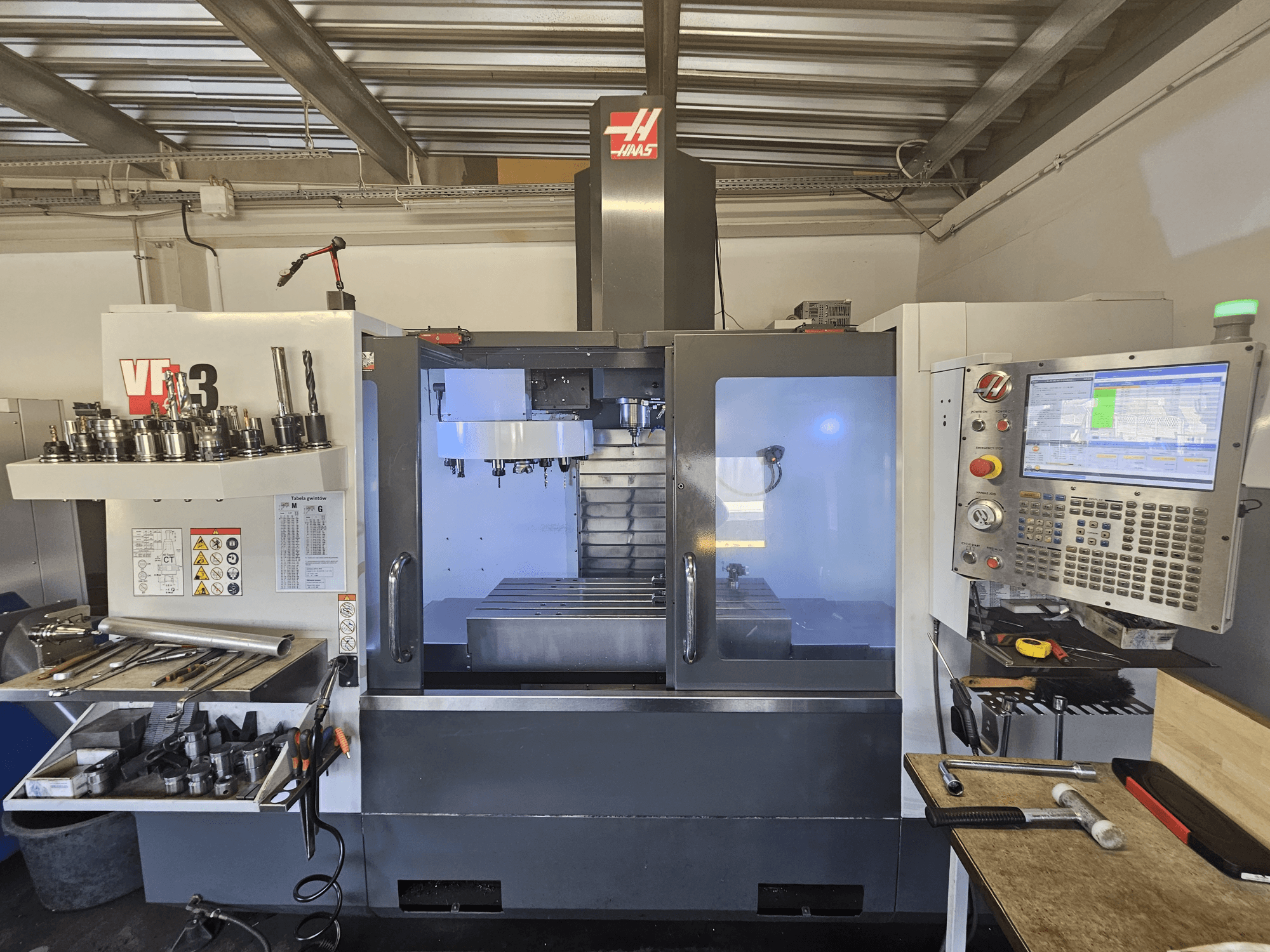 Haas VF-3 CNC staklės dirbtuvėse, vaizdas iš priekio, kuriame matomi įrankiai, valdikliai ir frezavimo zona su apšviestu vidumi.