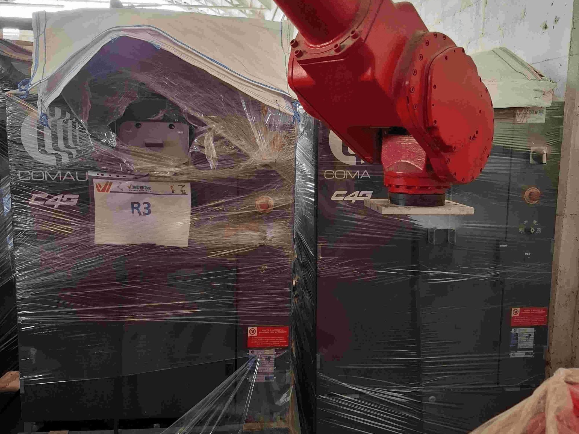 Iš priekio matoma "Comau C4G" roboto ranka, suvyniota į plastikinę plėvelę, su užrašu "R3", ant mašinos matomi įspėjamieji lipdukai.