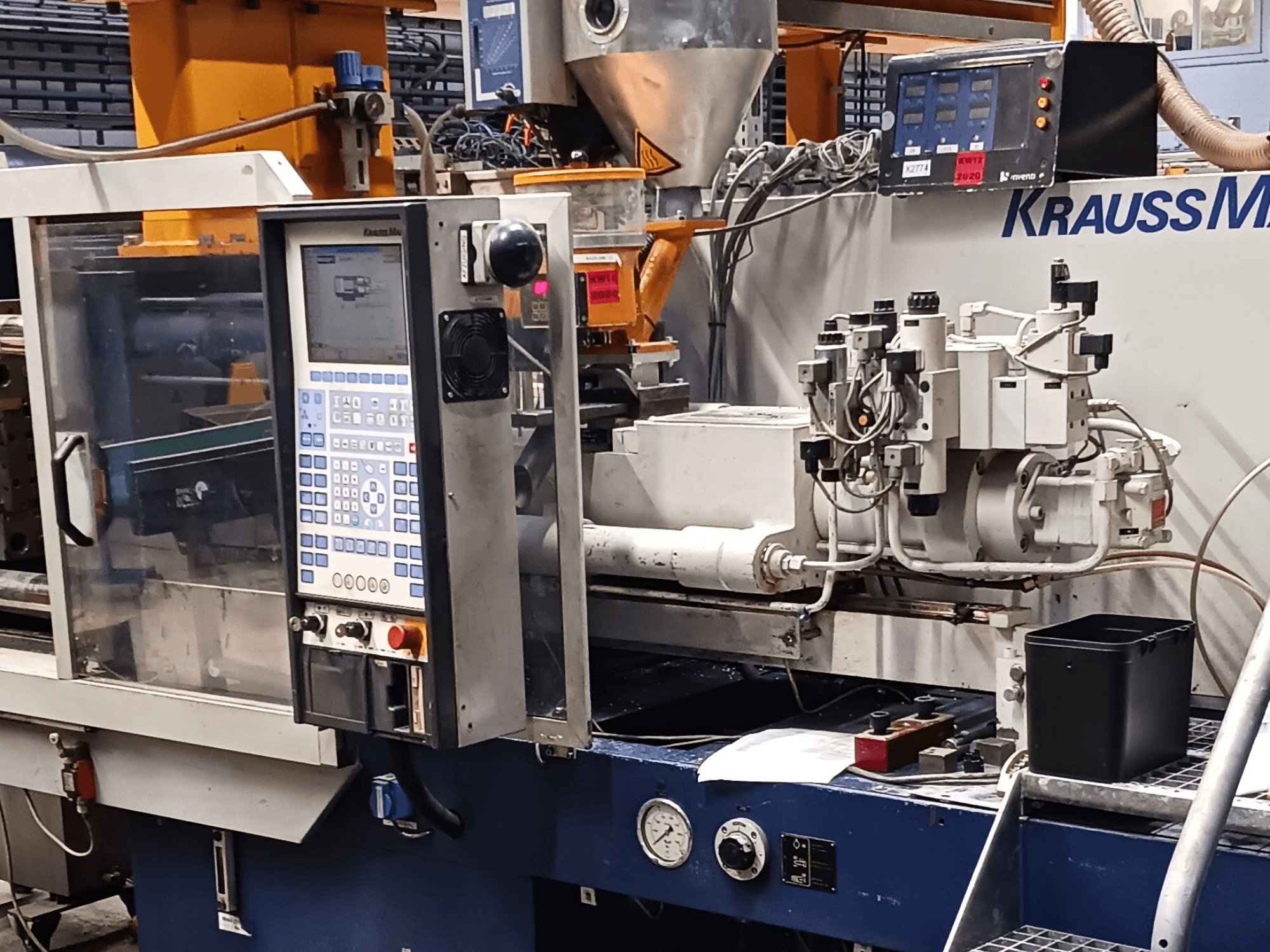 "KraussMaffei" liejimo mašina su valdymo pultu, hidrauliniais įrenginiais ir įrankiais pirmame plane, vaizdas iš priekio.