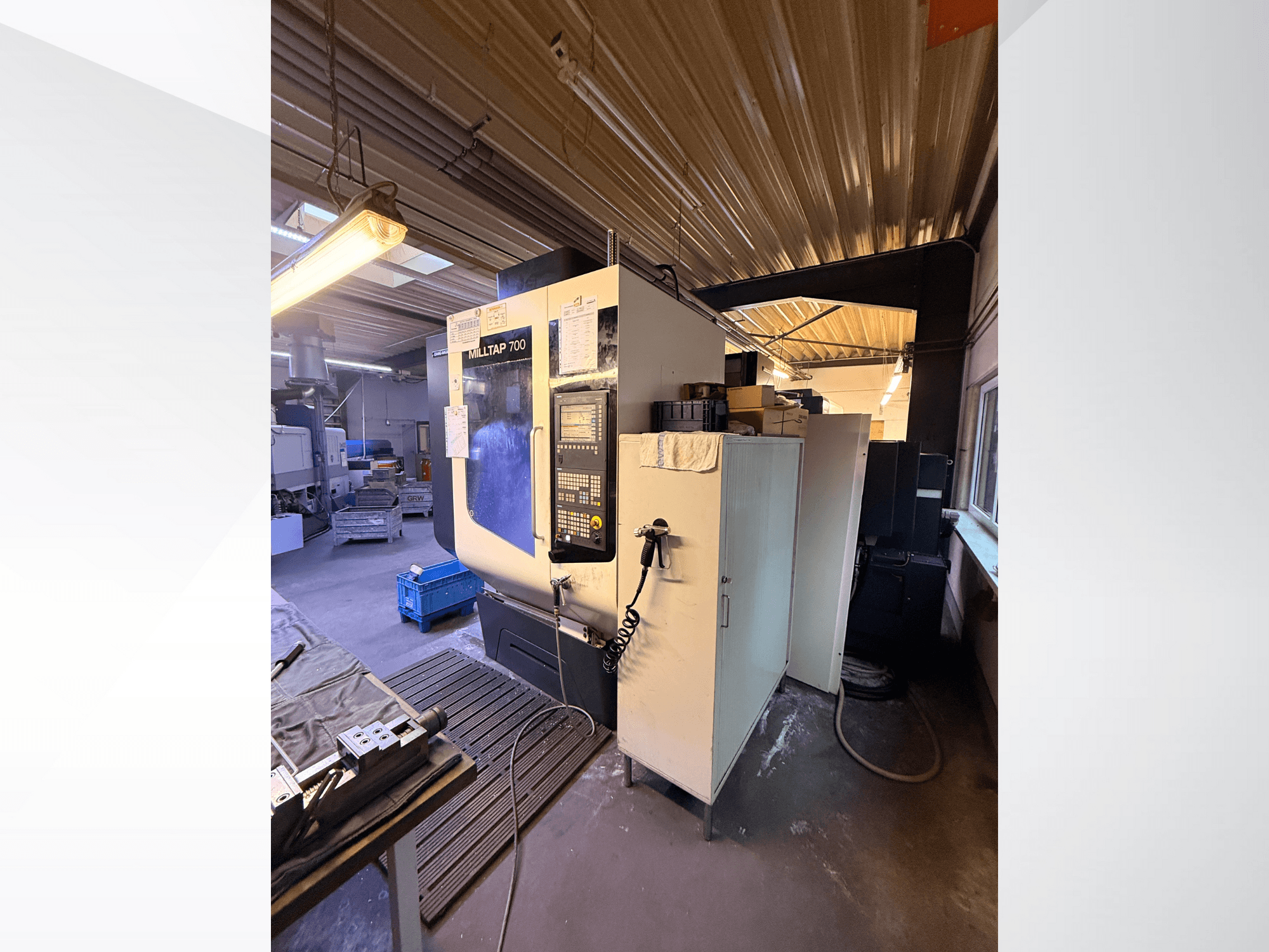 Milltap 700 CNC staklės dirbtuvėse; iš priekio matyti valdymo skydelis, įrankių lentynos ir laikymo vietos su apšvietimu virš jų.