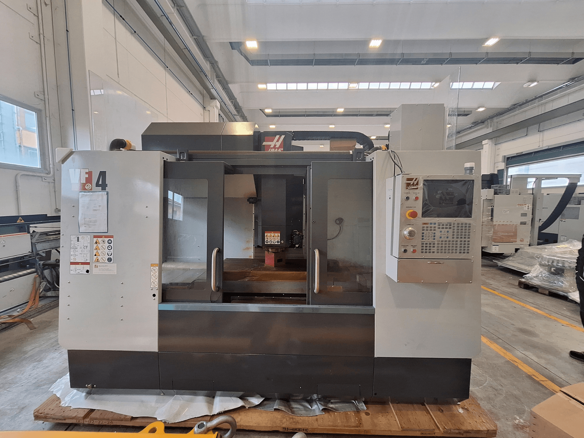 Vertikalusis apdirbimo centras „HAAS VF-4“, vaizdas iš priekio, kuriame matomas valdymo pultas, darbo zona ir saugos etiketės sandėlio aplinkoje.