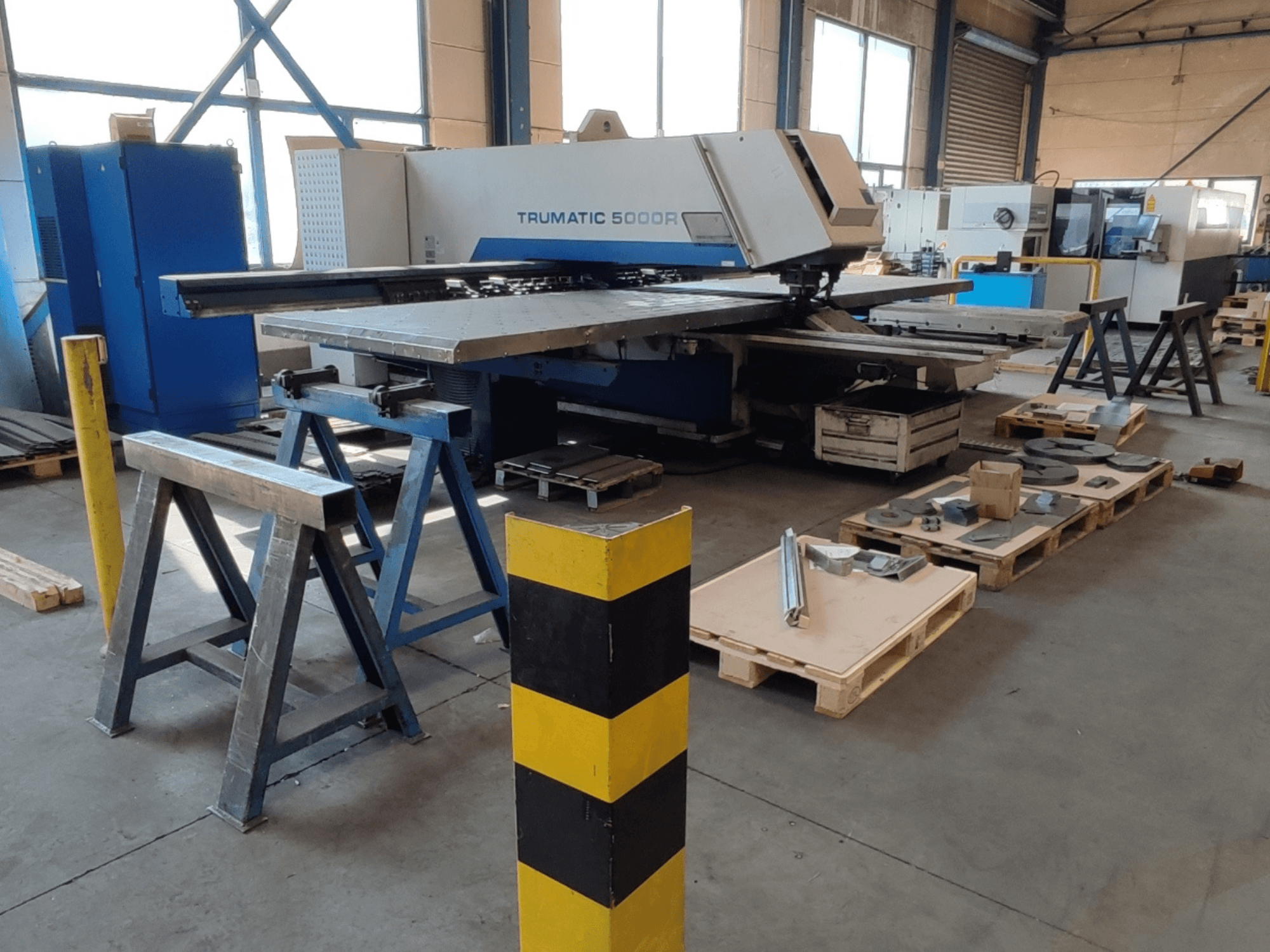 „TRUMPF Trumatic 5000 R“ CNC perforavimo staklės dirbtuvėse, vaizdas iš šono, šalia – ant padėklų sudėti įrankiai ir medžiagos.