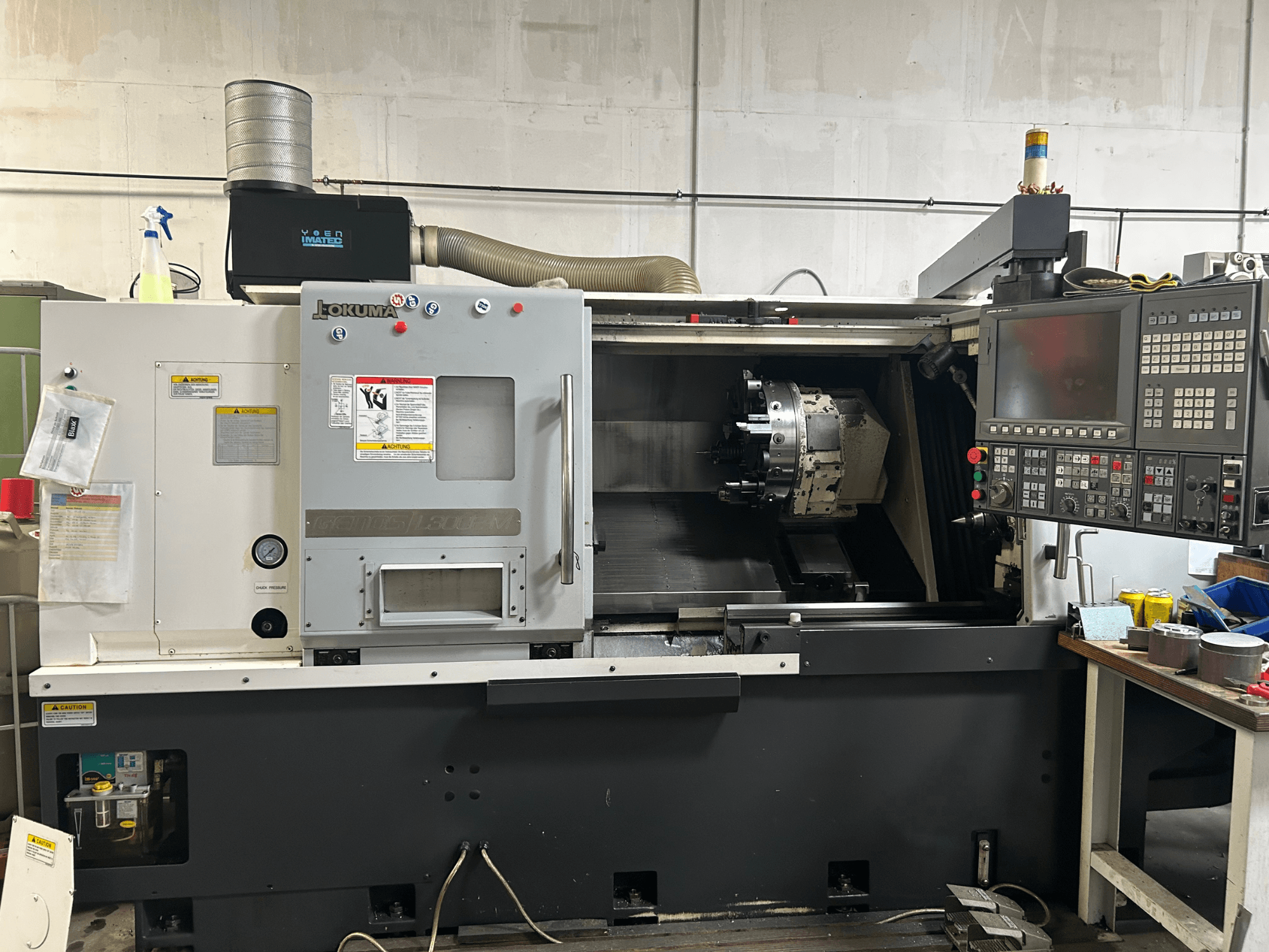 Horizontaliųjų tekinimo staklių "Okuma Genos L300E-M" vaizdas iš priekio su valdymo skydeliu, matomu sukliu ir įvairiais nustatymo įrankiais.