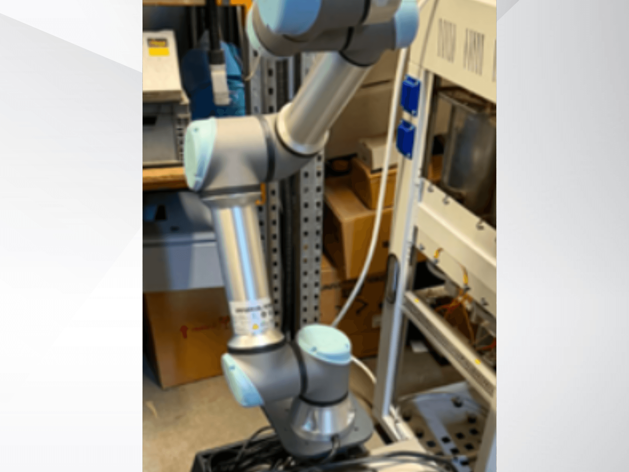 „Universal Robots“ UR5e robotinė ranka, matoma iš šono, su matomais lankstiniais sąnariais, lengva konstrukcija ir valdymo kabeliais.