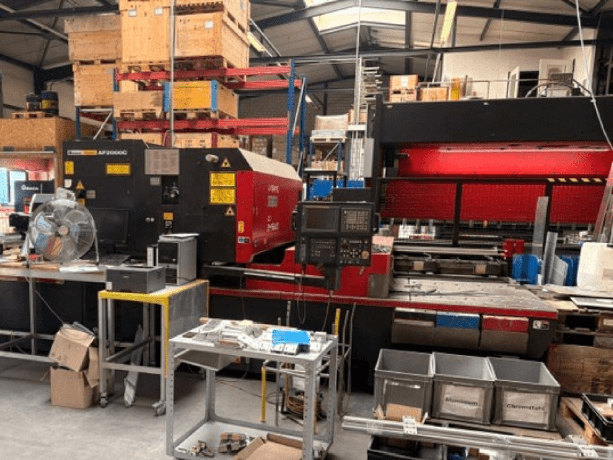 „AMADA LC-2415αIII“ CO₂ lazerio pjovimo staklės dirbtuvėse: matomas vaizdas iš priekio, valdymo pultas ir darbo erdvė.