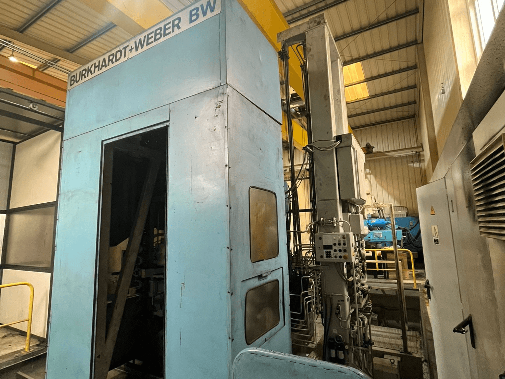 Burkhardt-Weber MC100 horizontalus apdirbimo centras, šoninis vaizdas, kuriame matomas mėlynas korpusas ir valdymo pultas su matomais vamzdeliais.