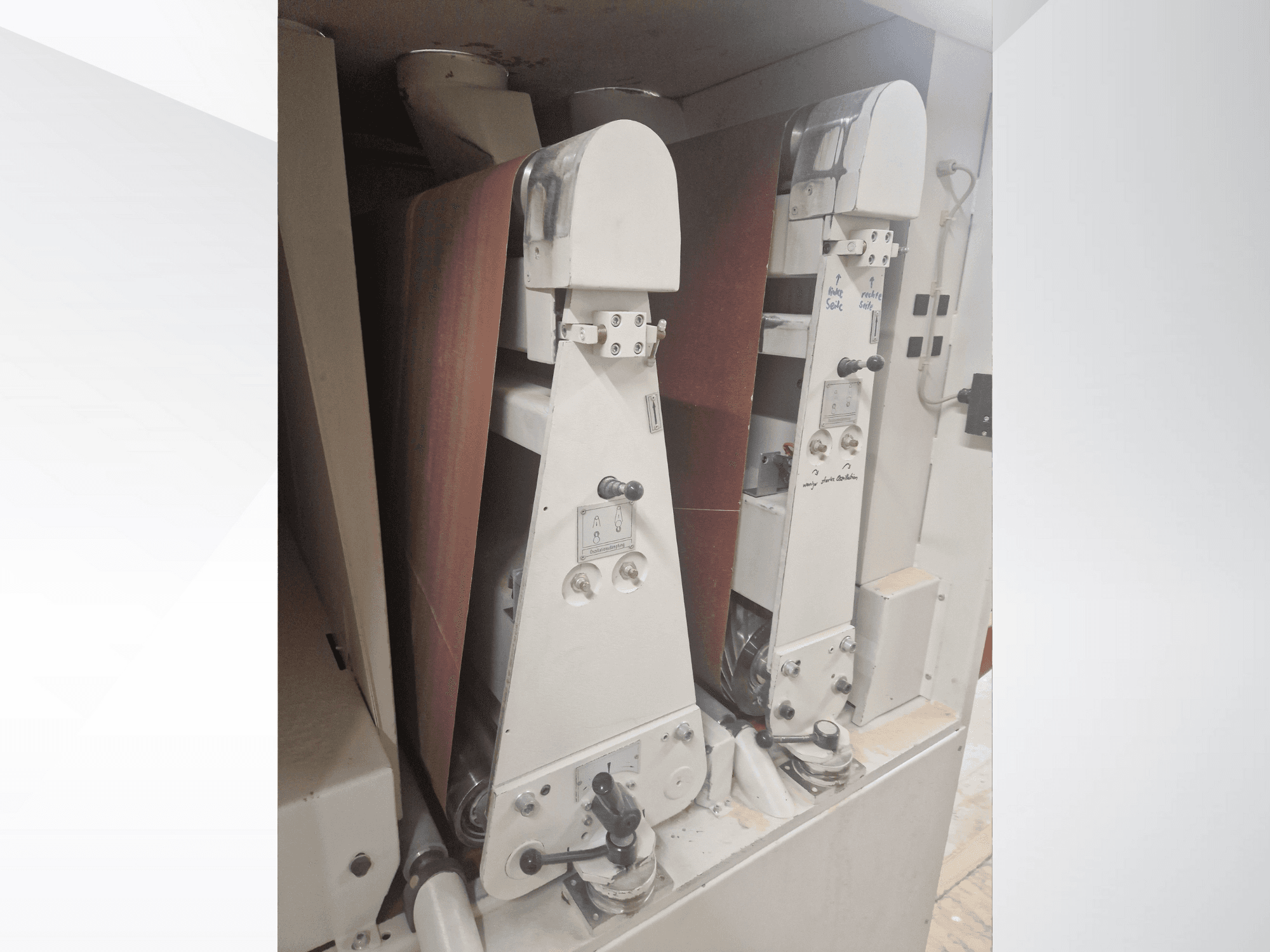 Dvi pramoninės šlifavimo mašinos su raudonomis šlifavimo juostomis, vaizdas iš priekio, matomi valdikliai ir metaliniai komponentai.