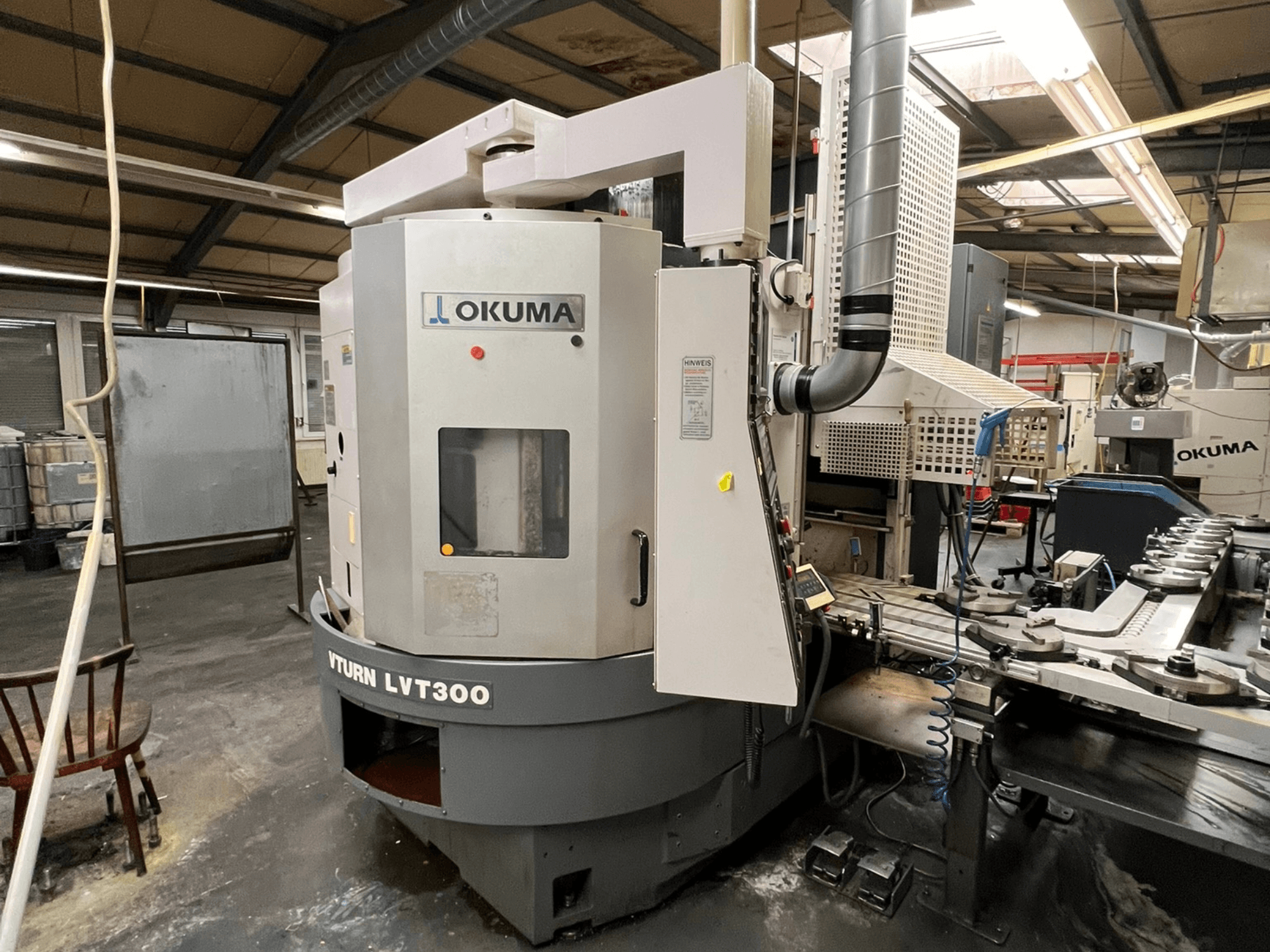 "Okuma Vturn LVT300" CNC tekinimo staklės, šoninis vaizdas pramoninėje aplinkoje, valdymo skydelis ir aušinimo skysčio sistema.