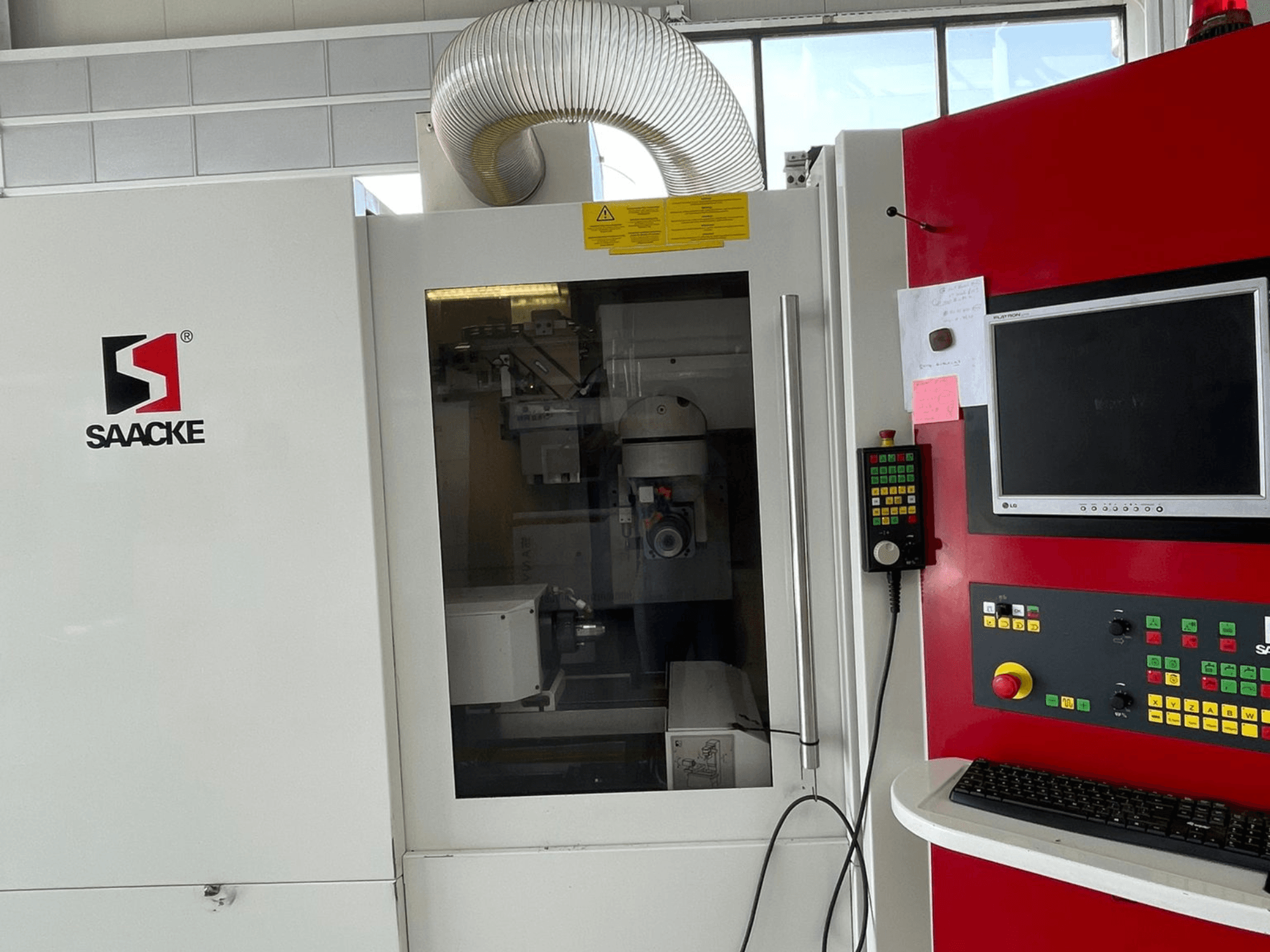 Baltos ir raudonos spalvos SAACKE mašina, vaizdas iš priekio, su valdymo pultu, monitoriumi ir per stiklą matomomis vidinėmis dalimis.