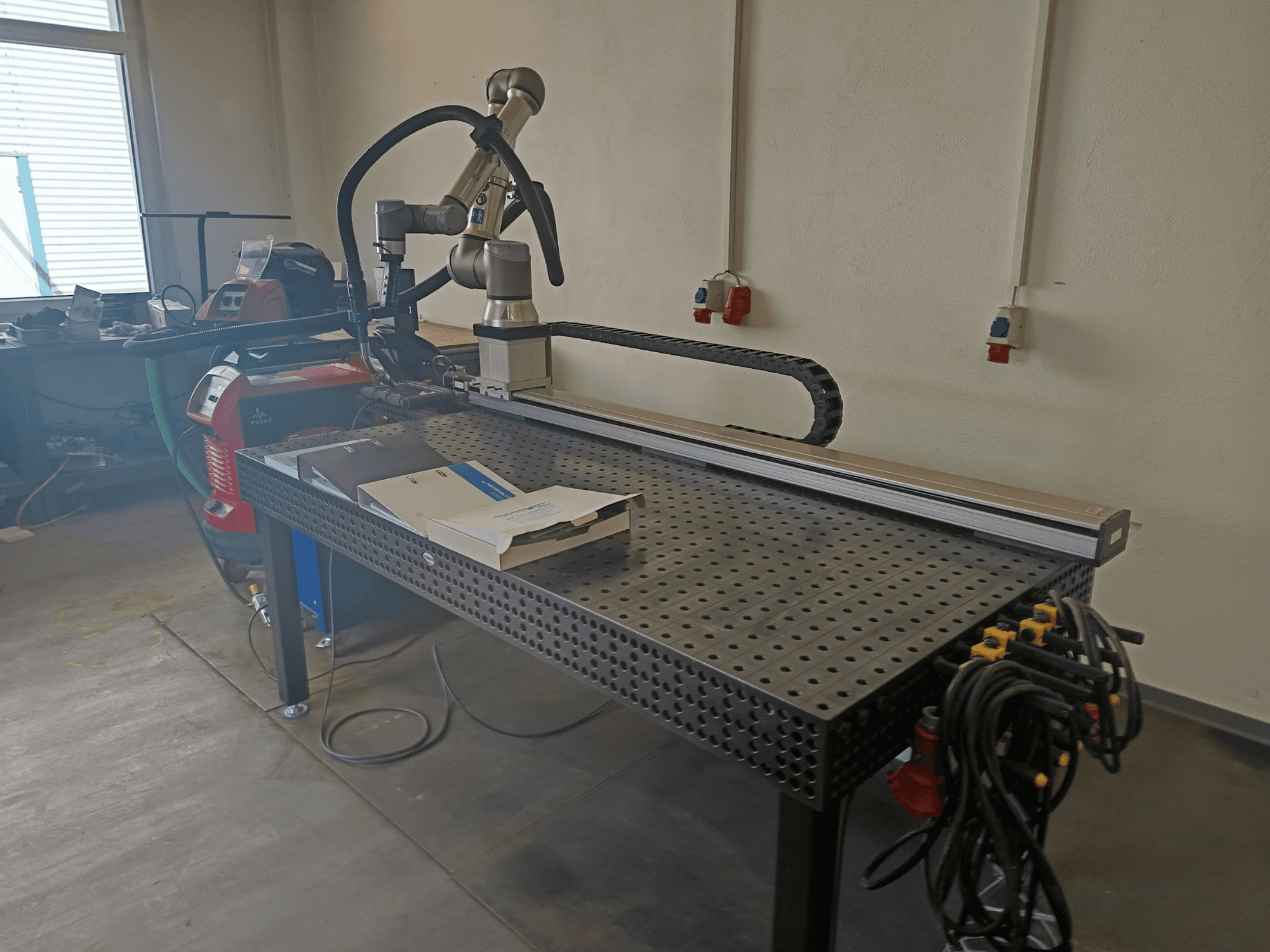 „FRONIUS TransSteel 4000“ impulsinio suvirinimo stendas, matomas iš šono, su robotine ranka, darbo zona ir maitinimo jungtimis.