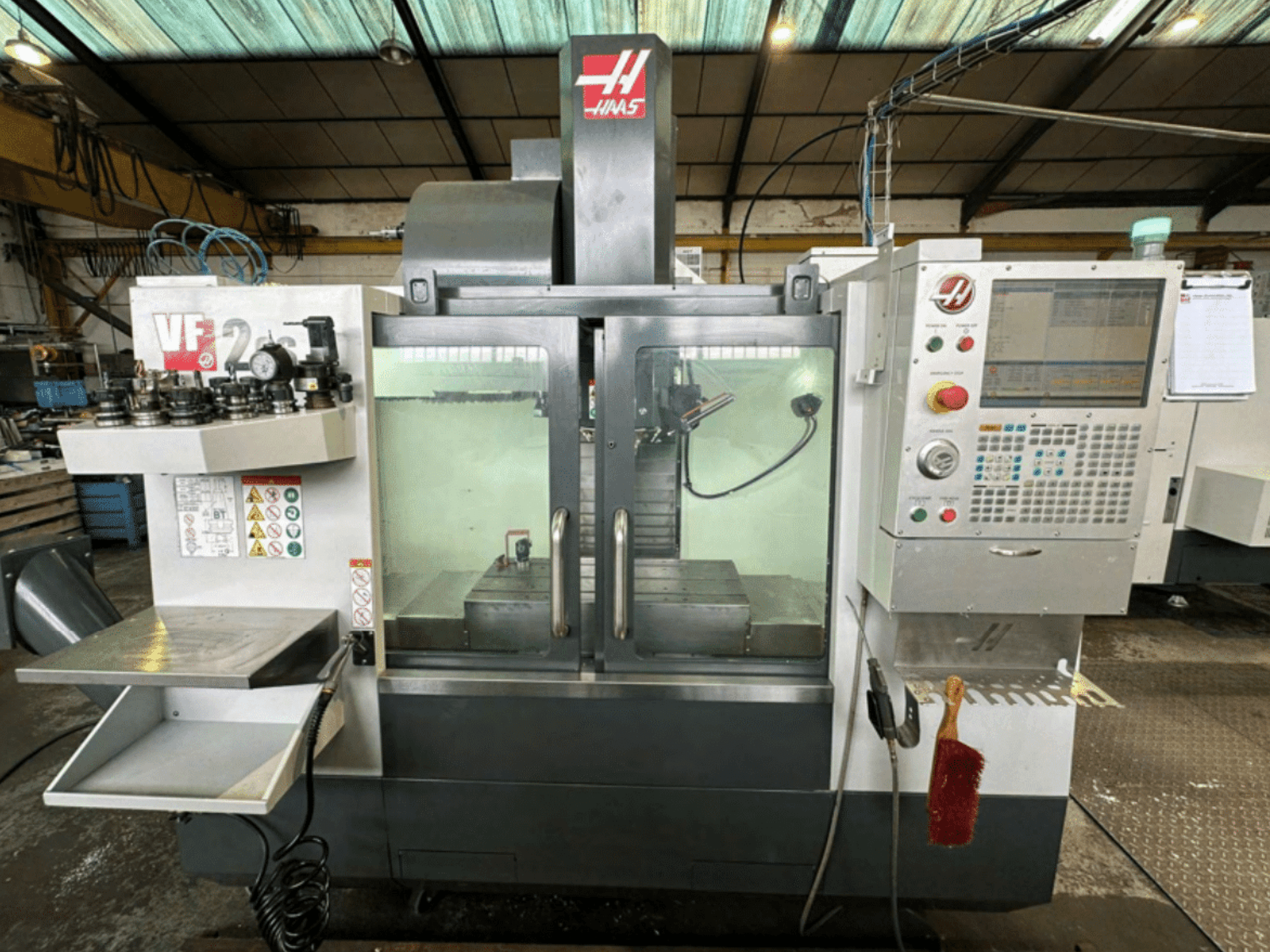 Haas VF2 CNC staklių priekinis vaizdas, kuriame matomas valdymo skydelis, įrankių laikikliai ir darbo vieta su saugos etiketėmis ir šepečiu.