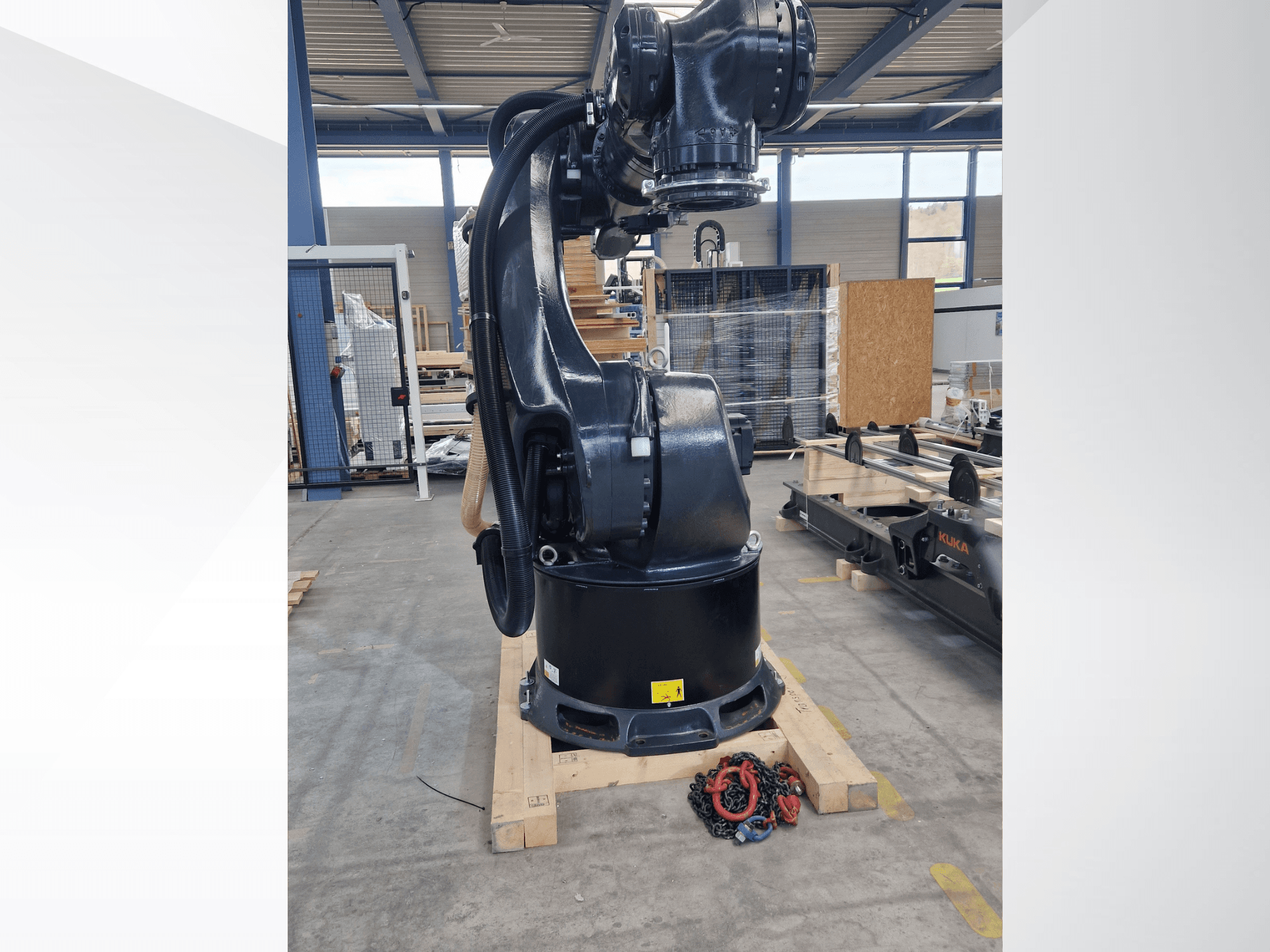 „KUKA KR 340 R3330“ roboto ranka sandėlyje, vaizdas iš šono, kuriame matoma jo bazė ir kabeliai, o pirmame plane – grandinės.