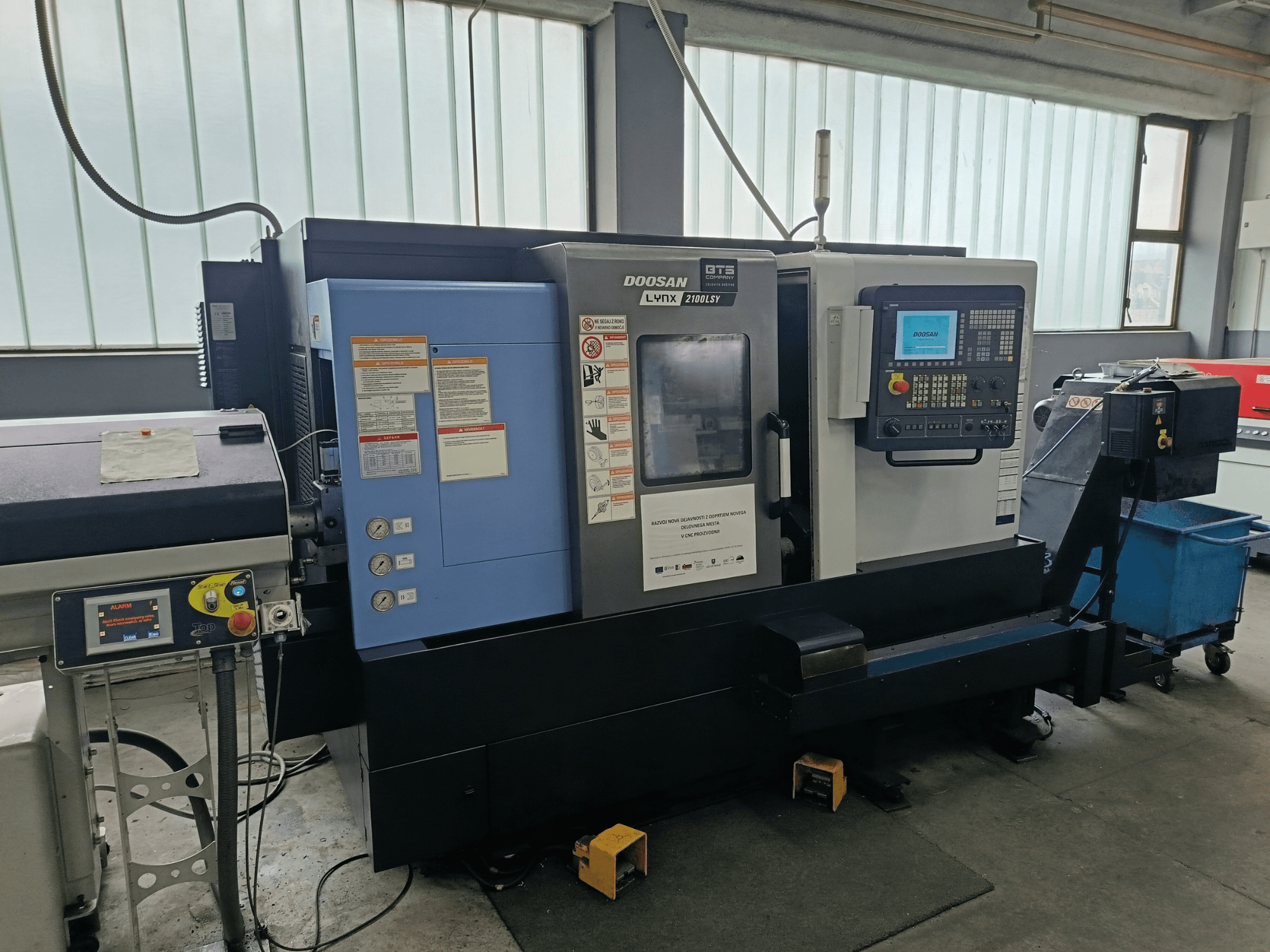 DOOSAN LYNX 2100 LSY tekinimo frezavimo centras, šoninis vaizdas, kuriame matomas valdymo skydelis ir valdymo mygtukai su informacinėmis etiketėmis.