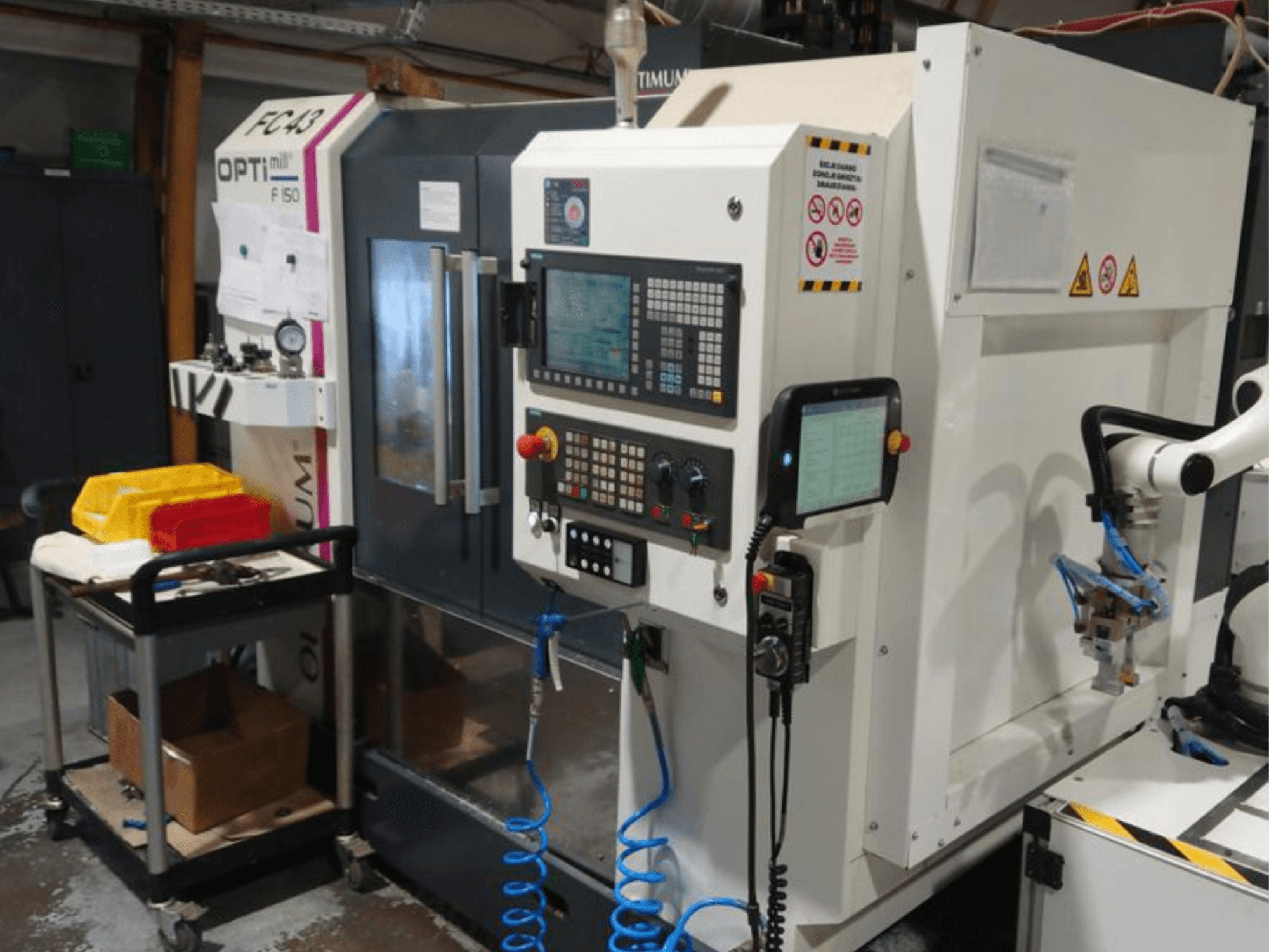 OPTIMUM F50 frezavimo staklių valdymo skydelis su žarnomis, įrankiais ir roboto ranka, matomas dirbtuvėse.